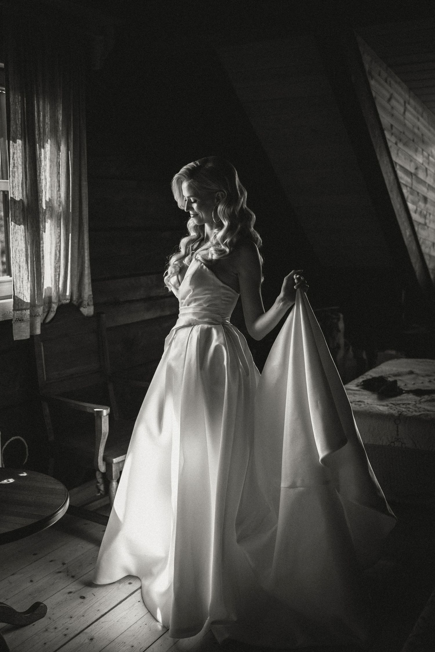 Black and white photo of a wedding dress silhouette standing by a window in rustic setting.