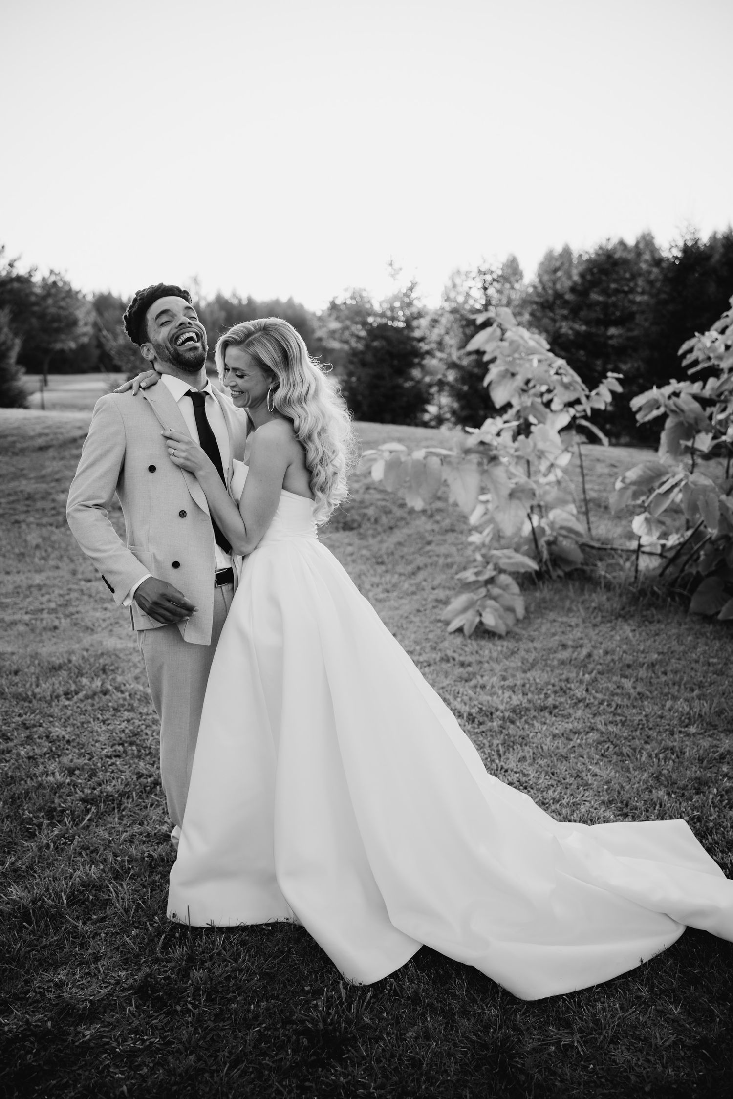 Black and white photo of a bride in flowing wedding gown and groom in light suit laughing together outdoors.