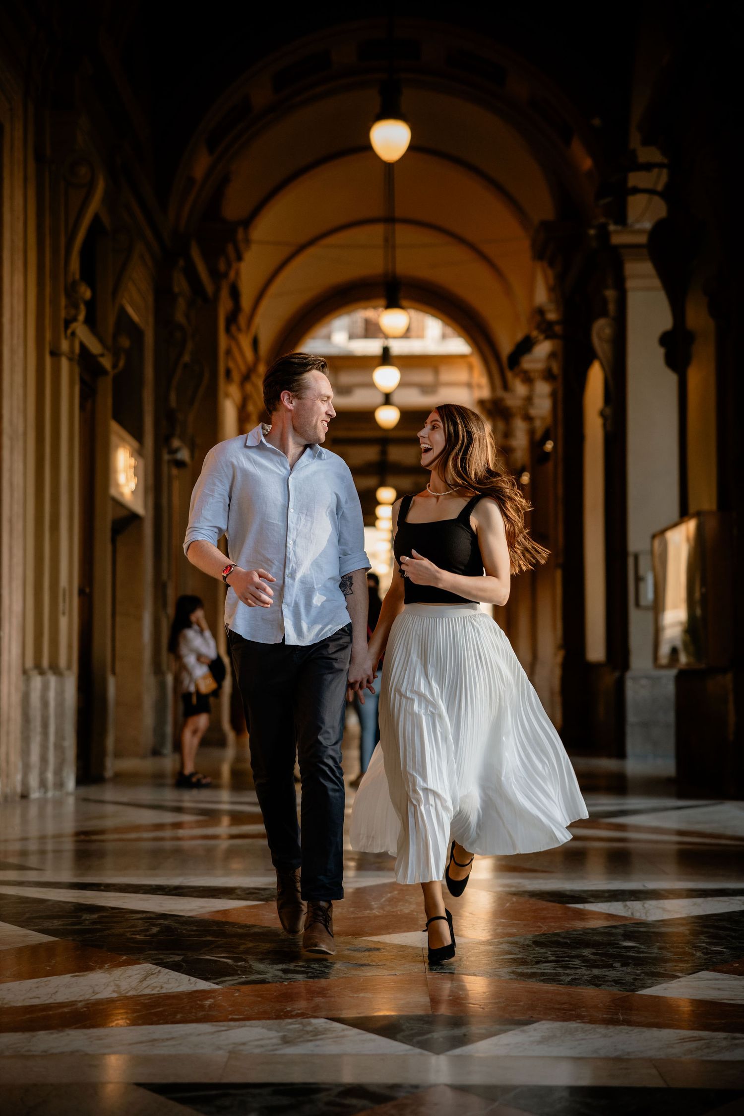 A couple walks together under ornate arches in an elegant historic corridor with warm lighting and marble floors.