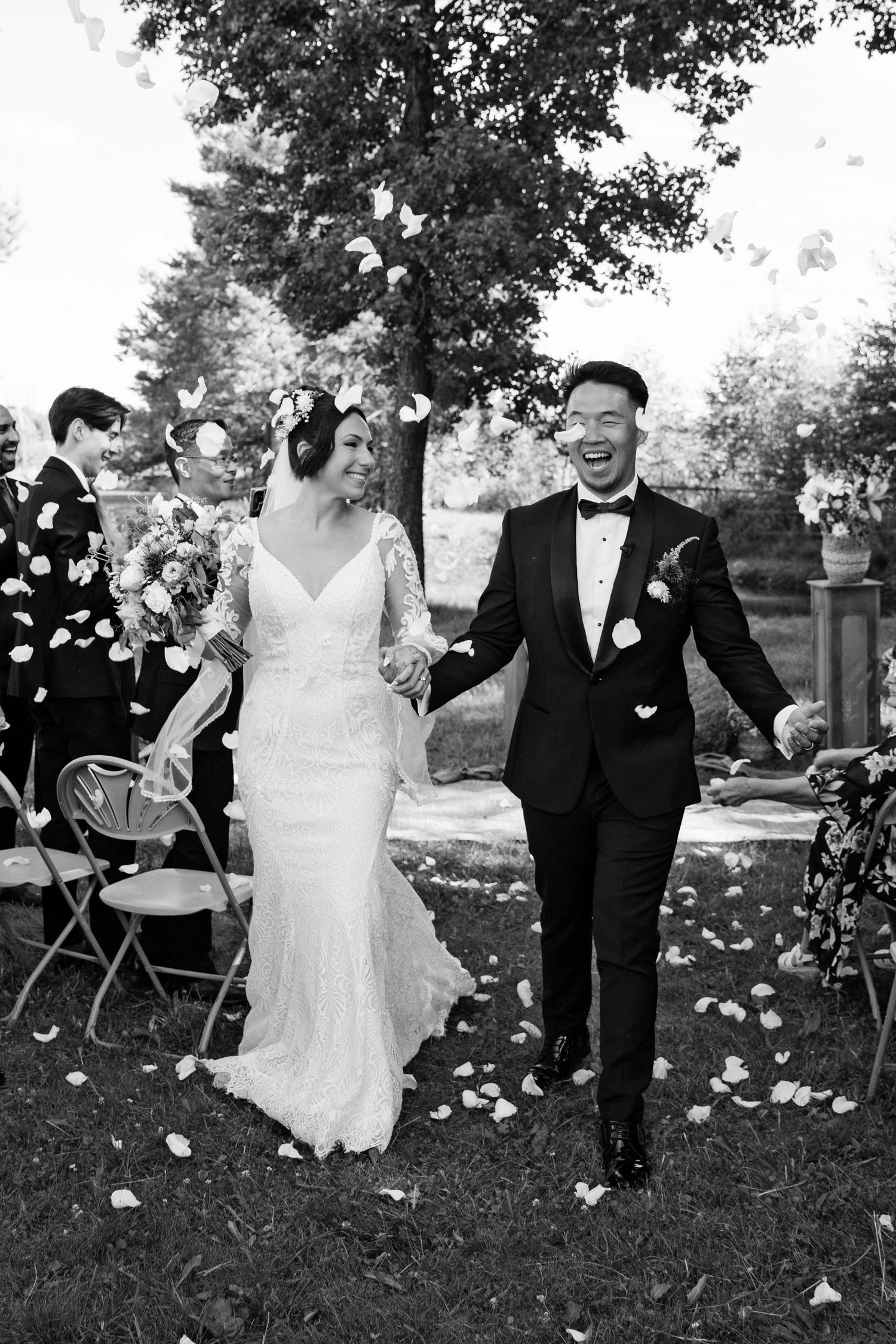 A black and white wedding photograph capturing a joyful couple walking through flower petals on their special day.