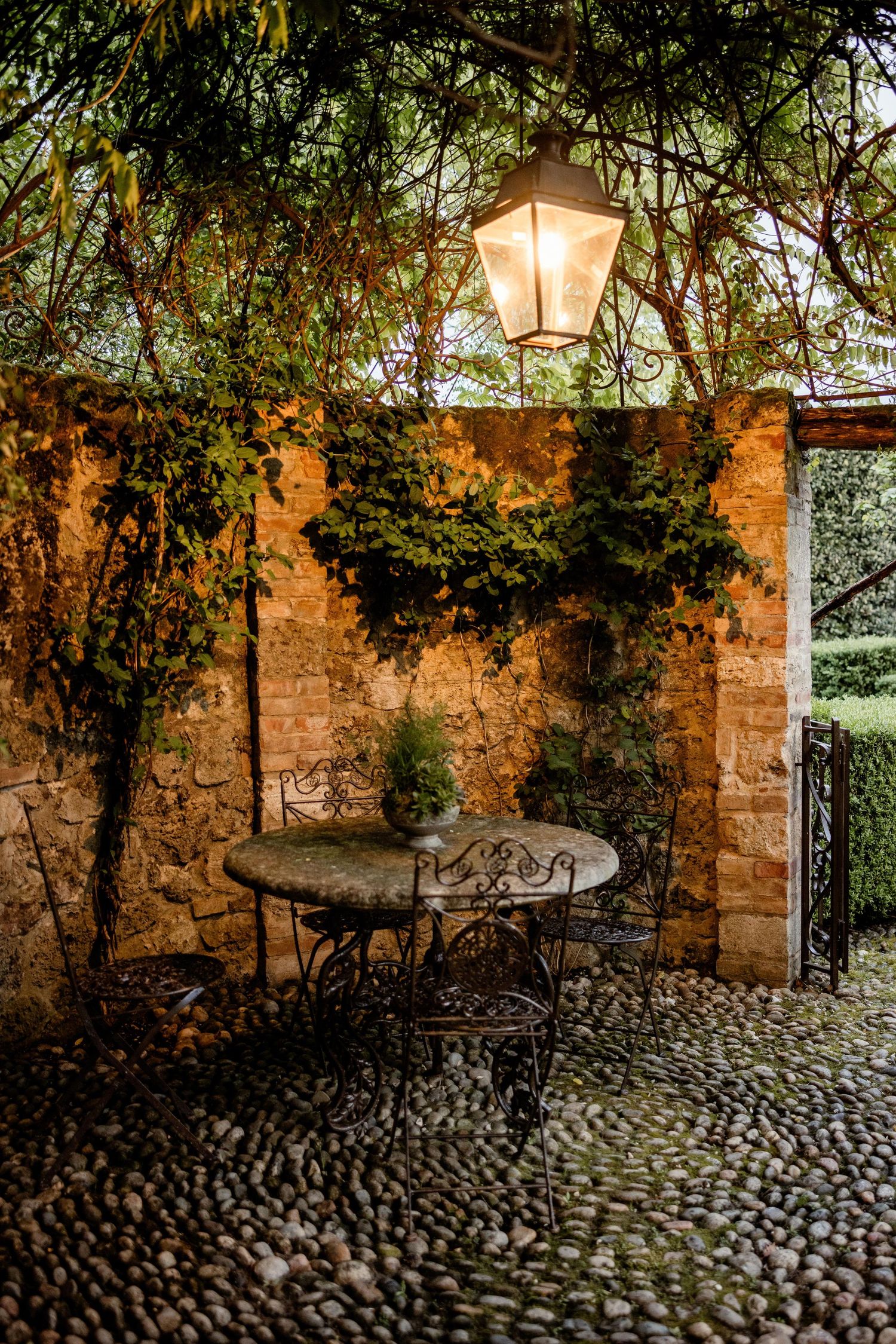 Rustic stone patio with wrought iron table and chairs under glowing lantern light with ivy-covered walls at dusk.