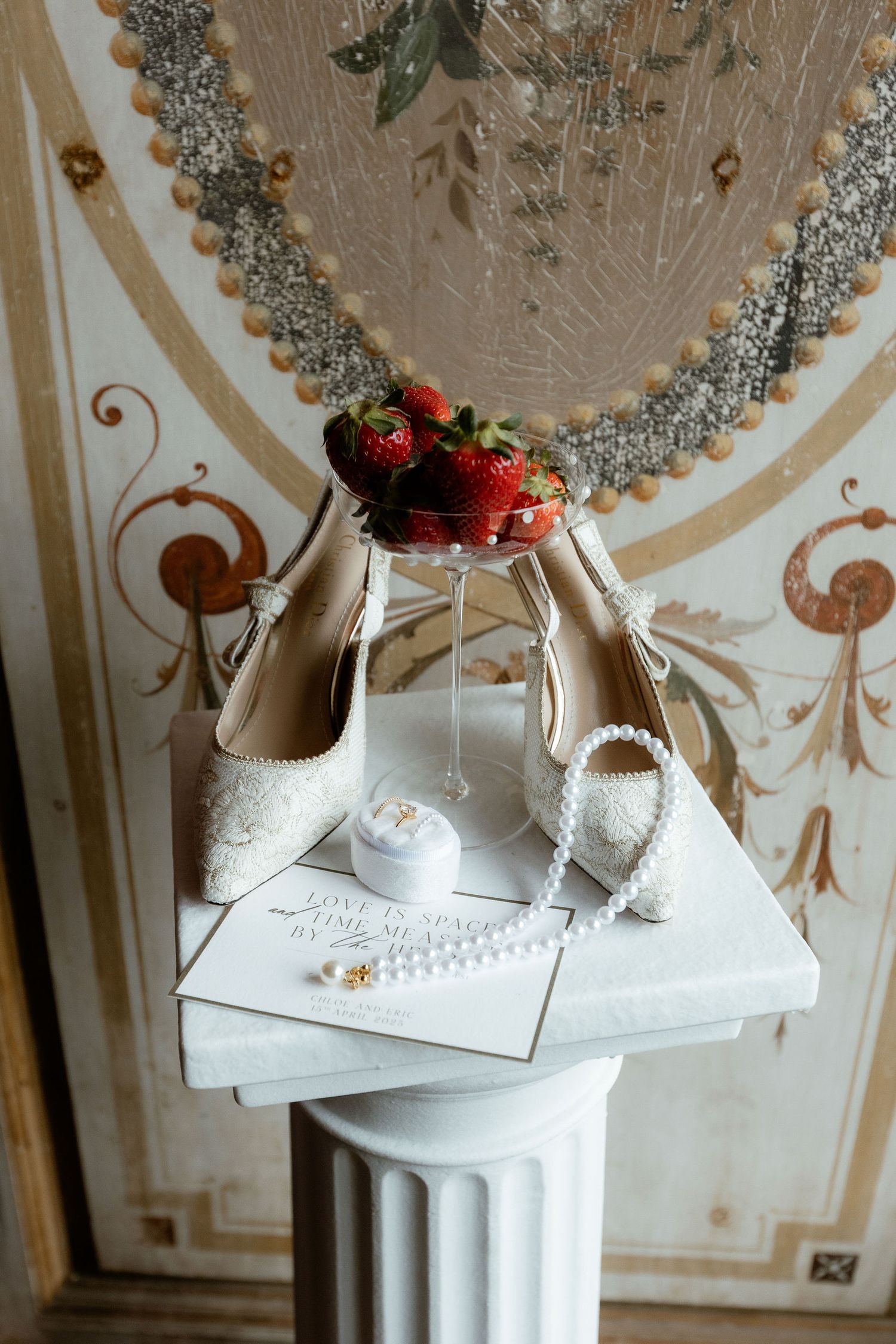 Elegant white bridal shoes and rings displayed on a classical column pedestal with roses and silver decor in the background.