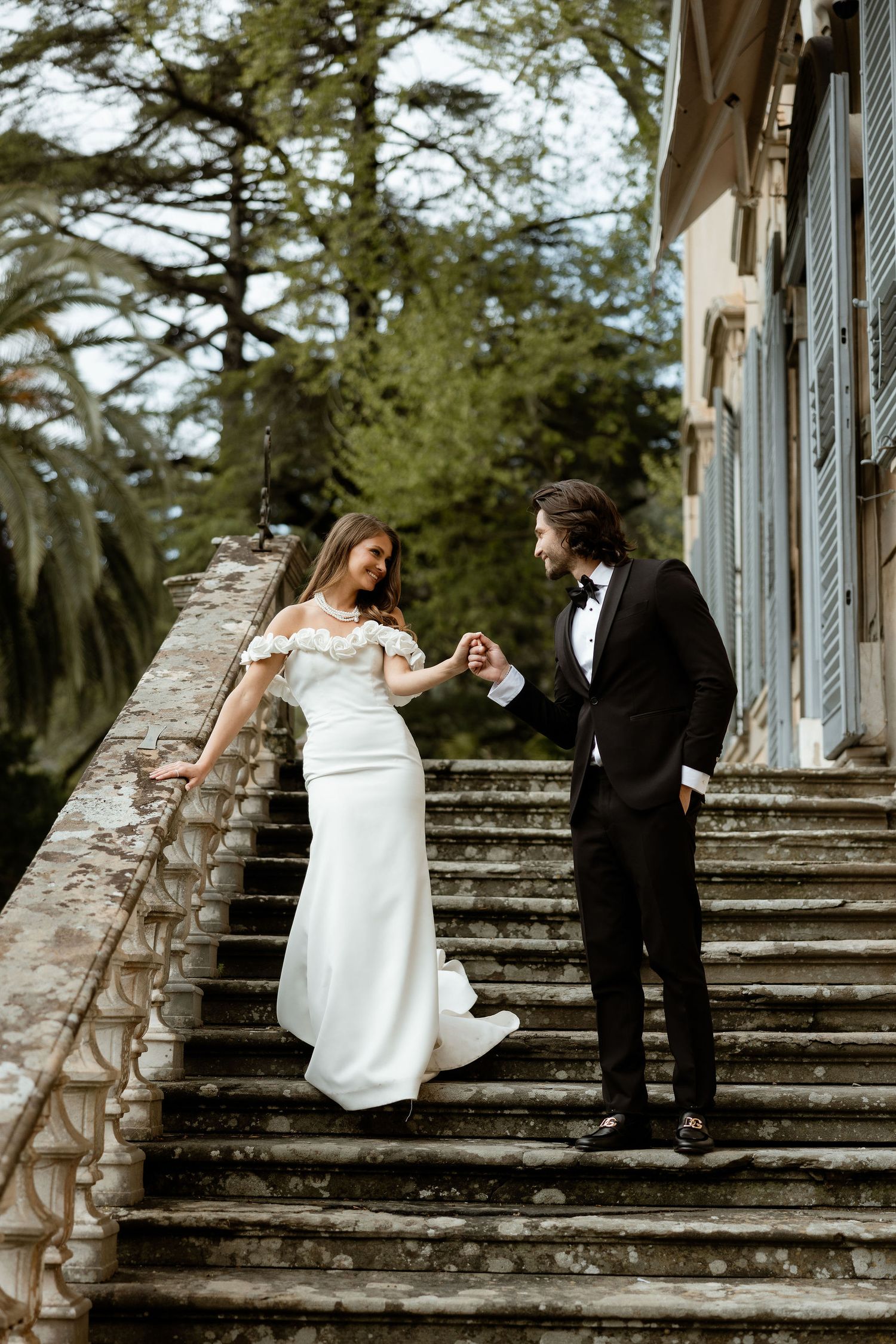 A couple in wedding attire stands on an elegant stone staircase surrounded by lush greenery at a historic venue.