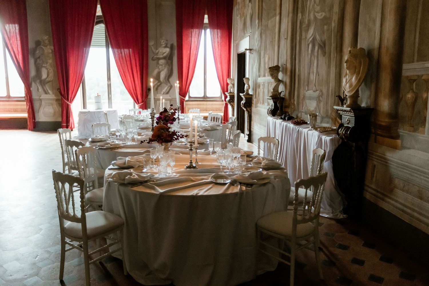Elegant dining room with round table set with white linens, glassware and red curtains in an ornate historic interior.