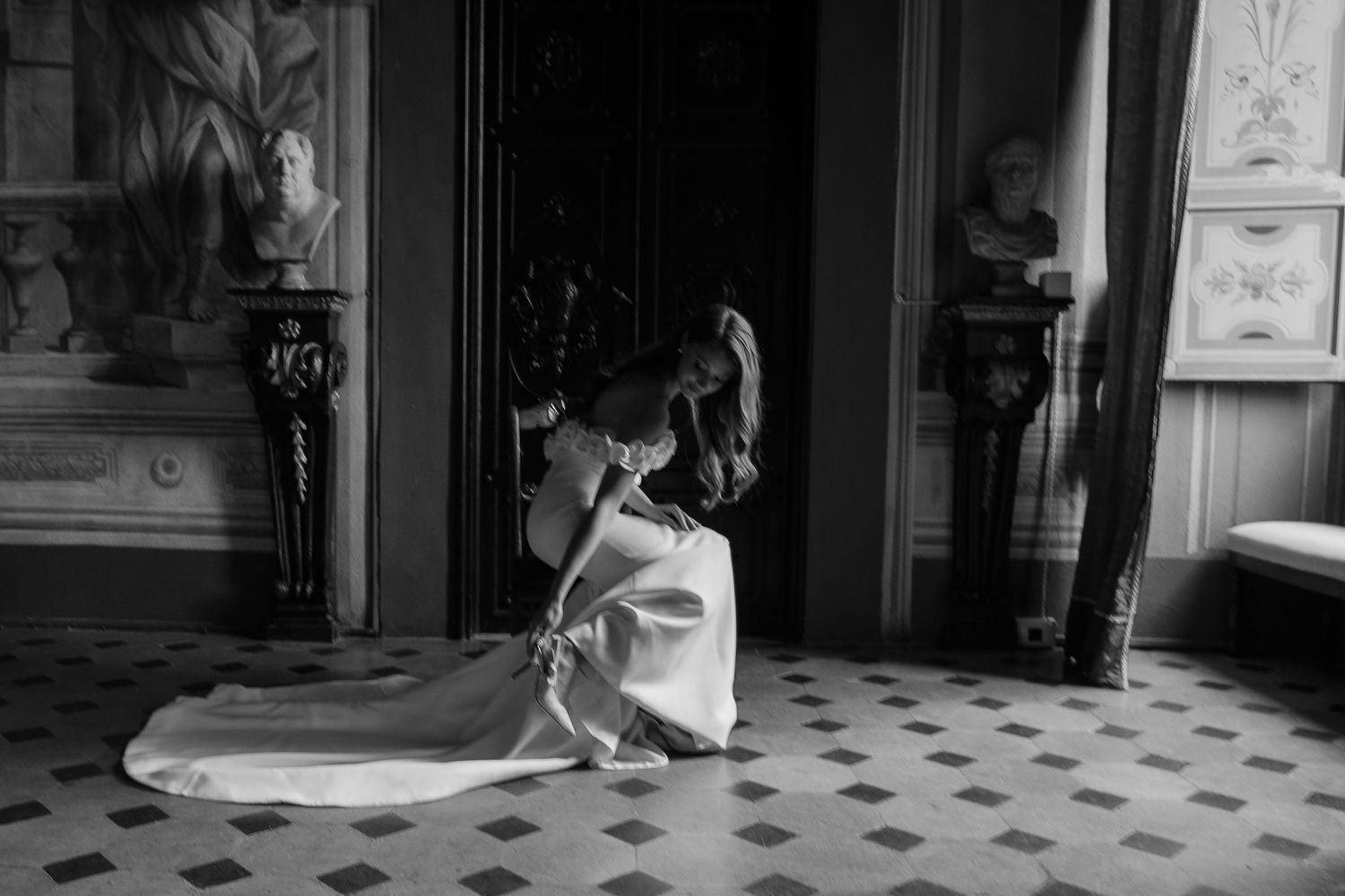 In a grand ornate room with tiled floors, a person in flowing white dress kneels elegantly beside a window.