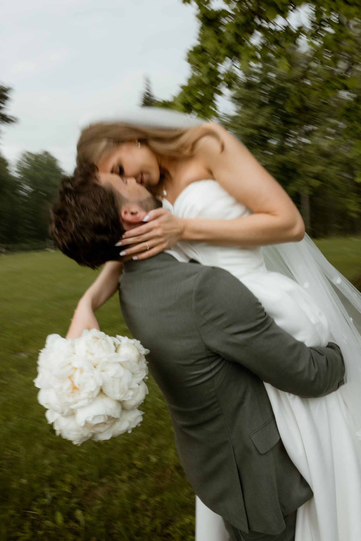 A romantic wedding couple embraces and kisses while holding a white peony bouquet in an outdoor garden setting.