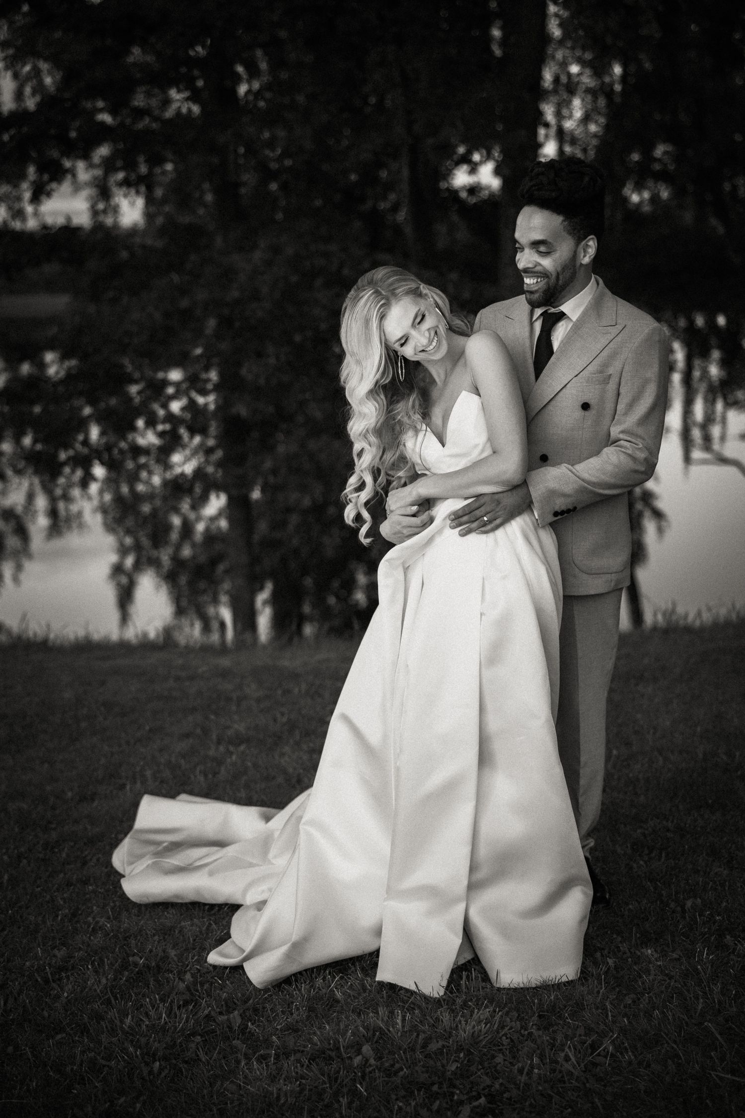 A romantic black and white wedding photo of a couple embracing on a grassy lawn near trees.