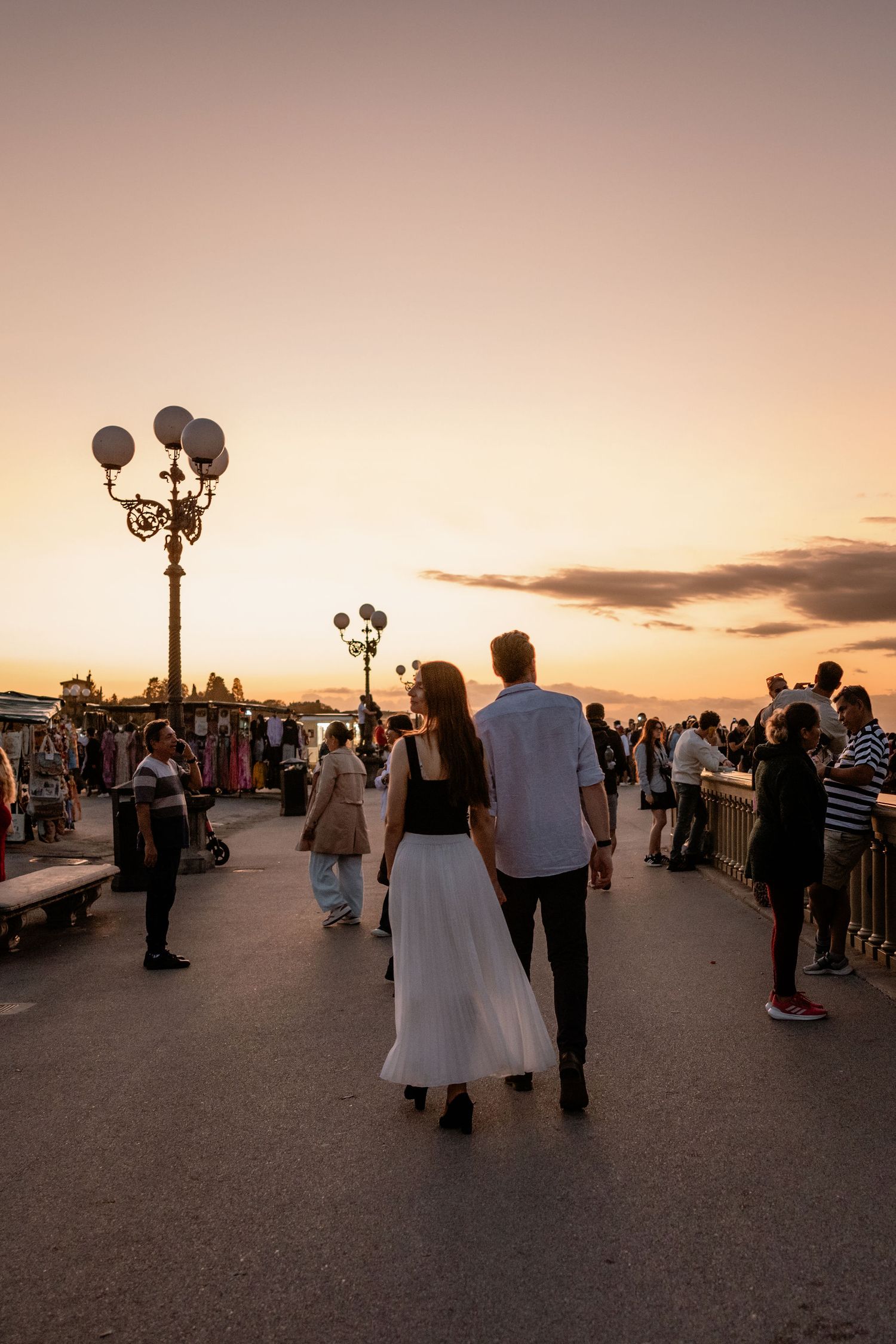 Crowds gather at a park during sunset with decorative lampposts and people strolling along the promenade.