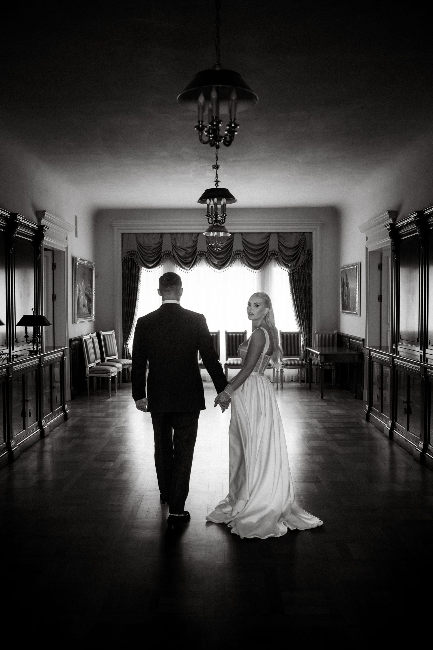 A black and white photo shows a couple walking hand in hand down an elegant hallway.