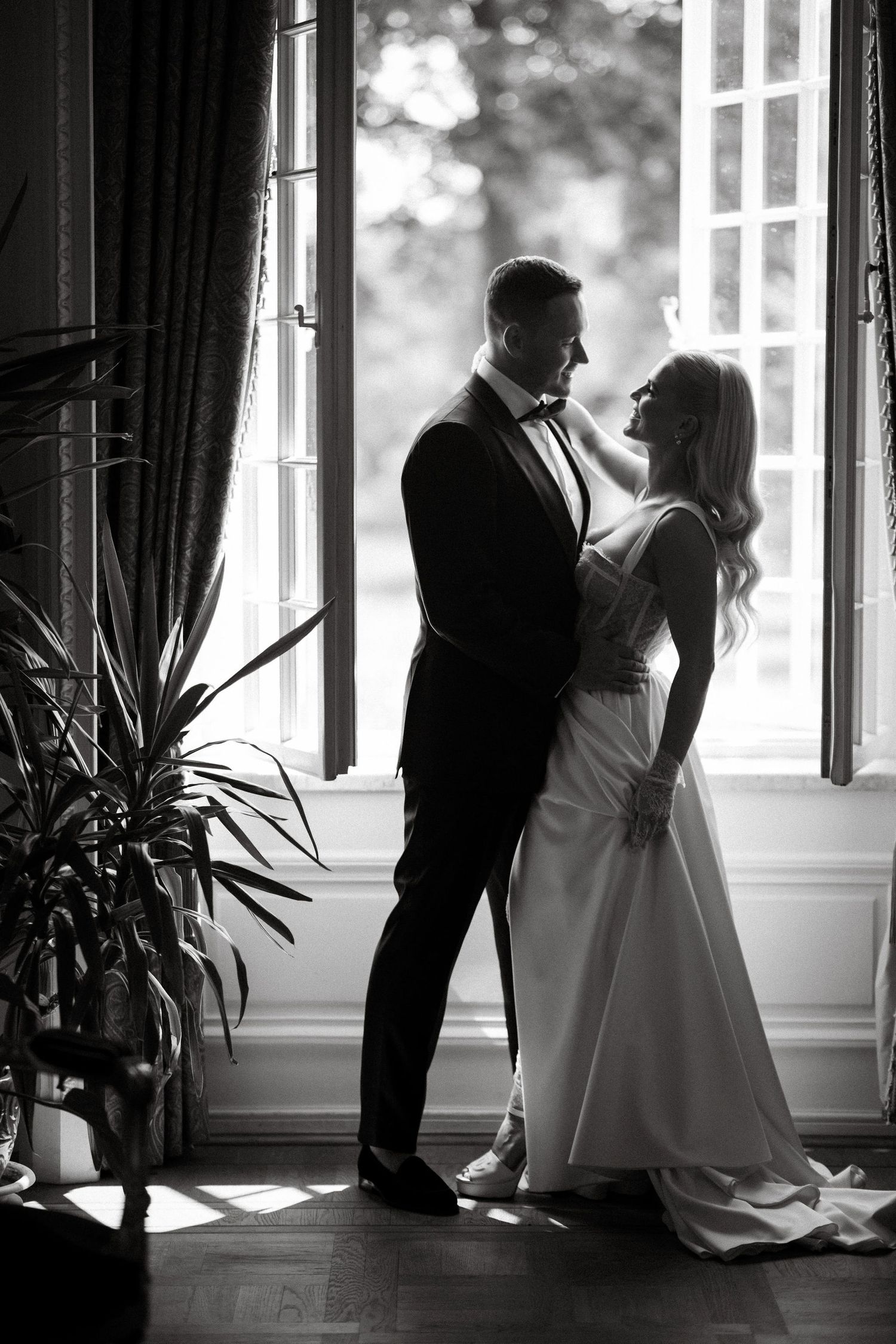 Elegant silhouette of a couple in formal attire standing by large windows in romantic black and white photography.