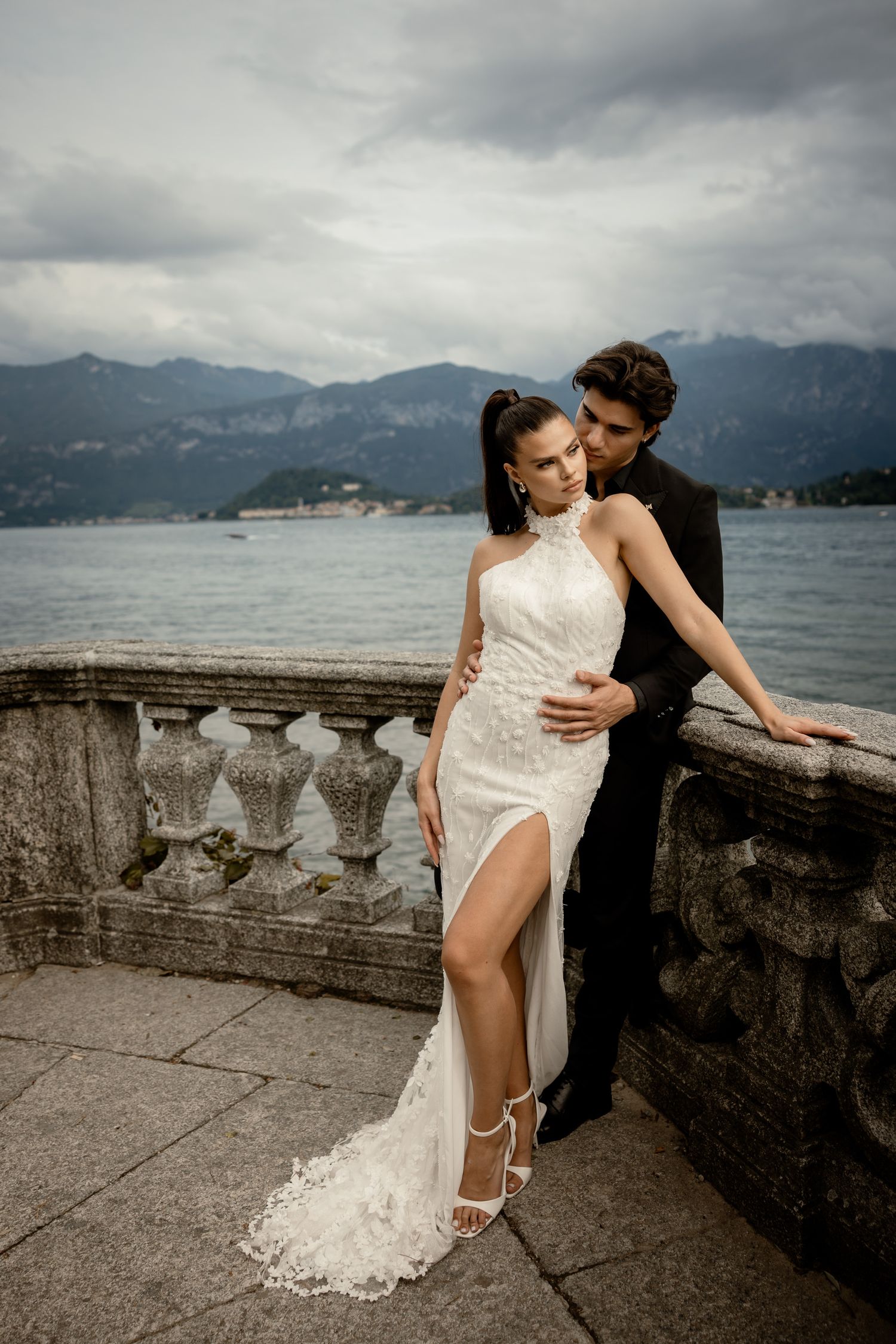 Bride in elegant white dress with slit poses with groom on lakeside terrace with mountain views.