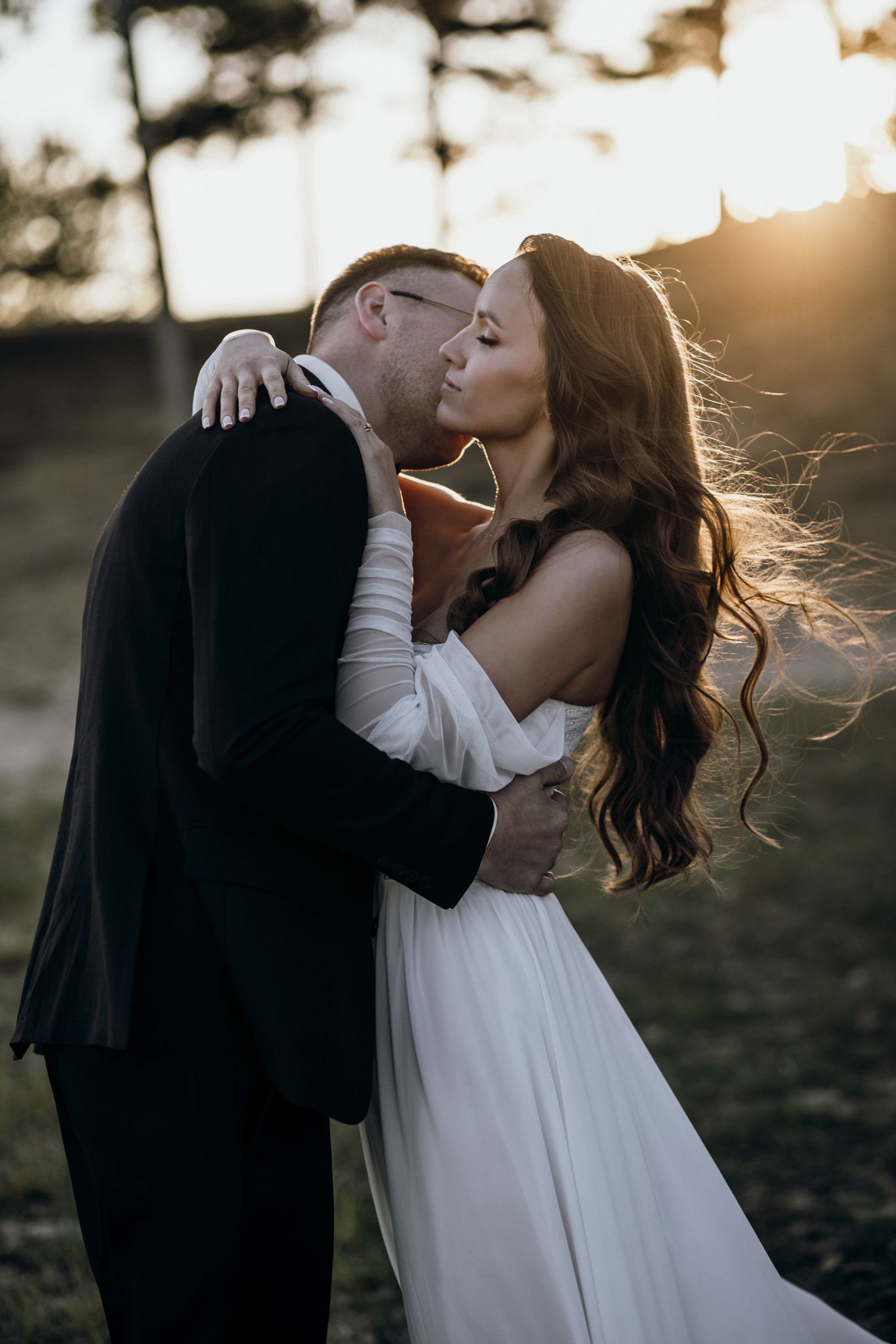 A romantic couple shares a tender kiss at sunset while wearing formal wedding attire in a dreamy outdoor setting.