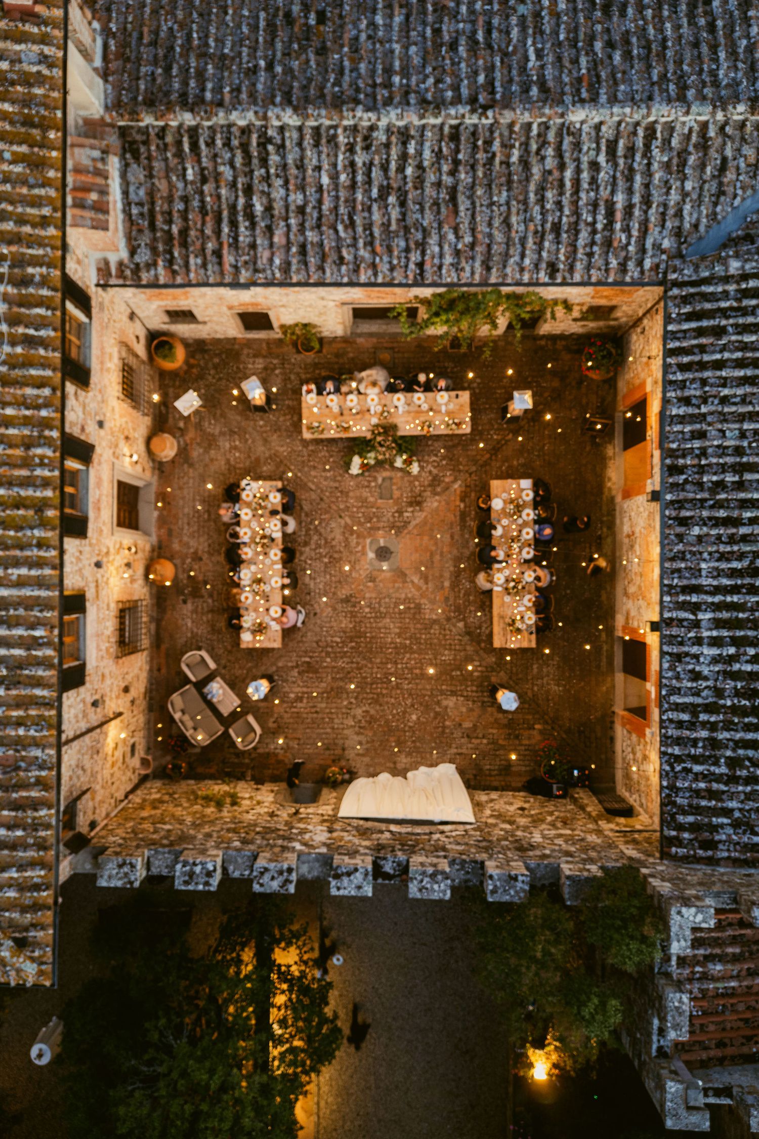 Aerial view of an outdoor wedding reception venue at night with long tables and string lights in a brick courtyard.