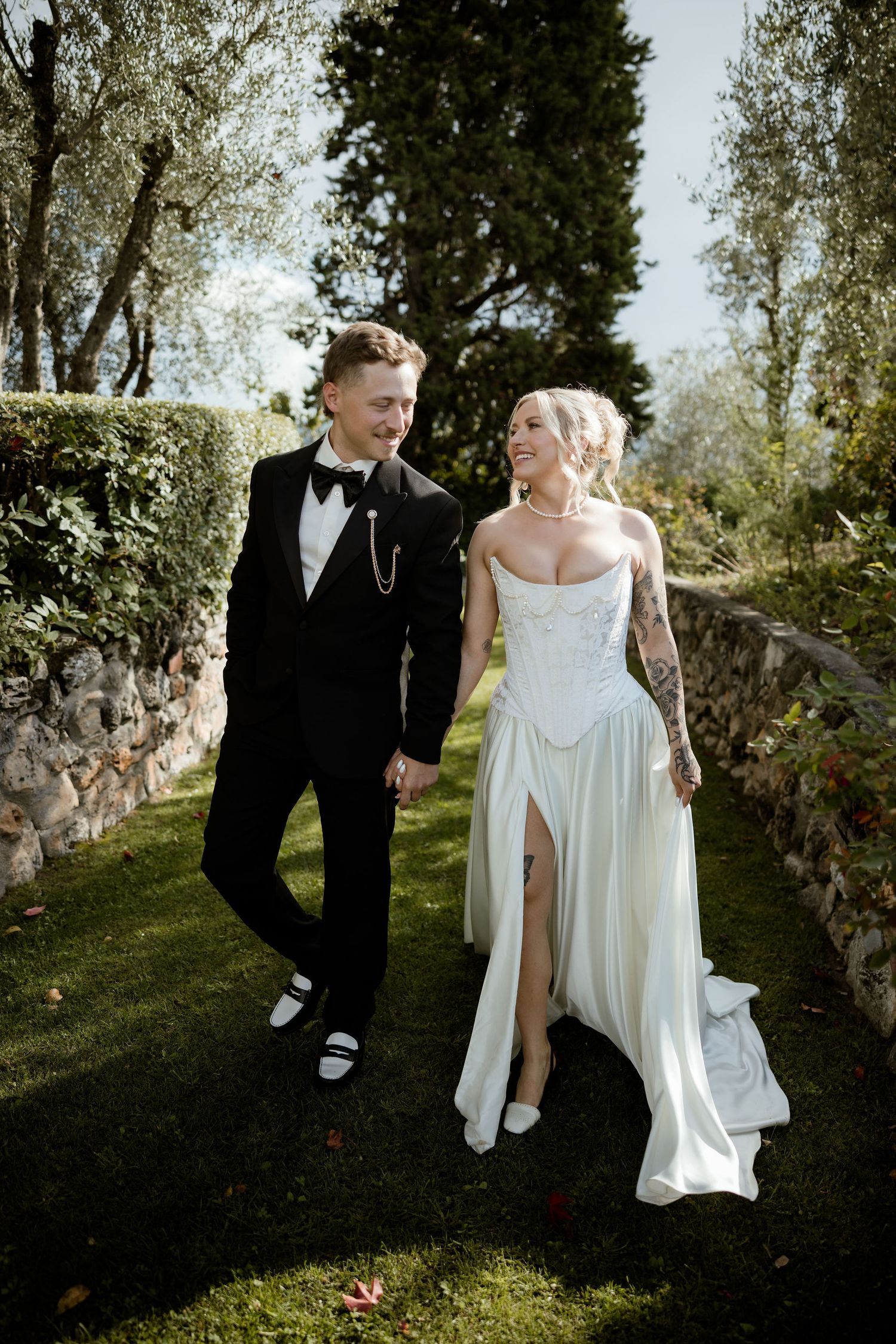 A couple in wedding attire walks together through an olive grove on a sunny afternoon.