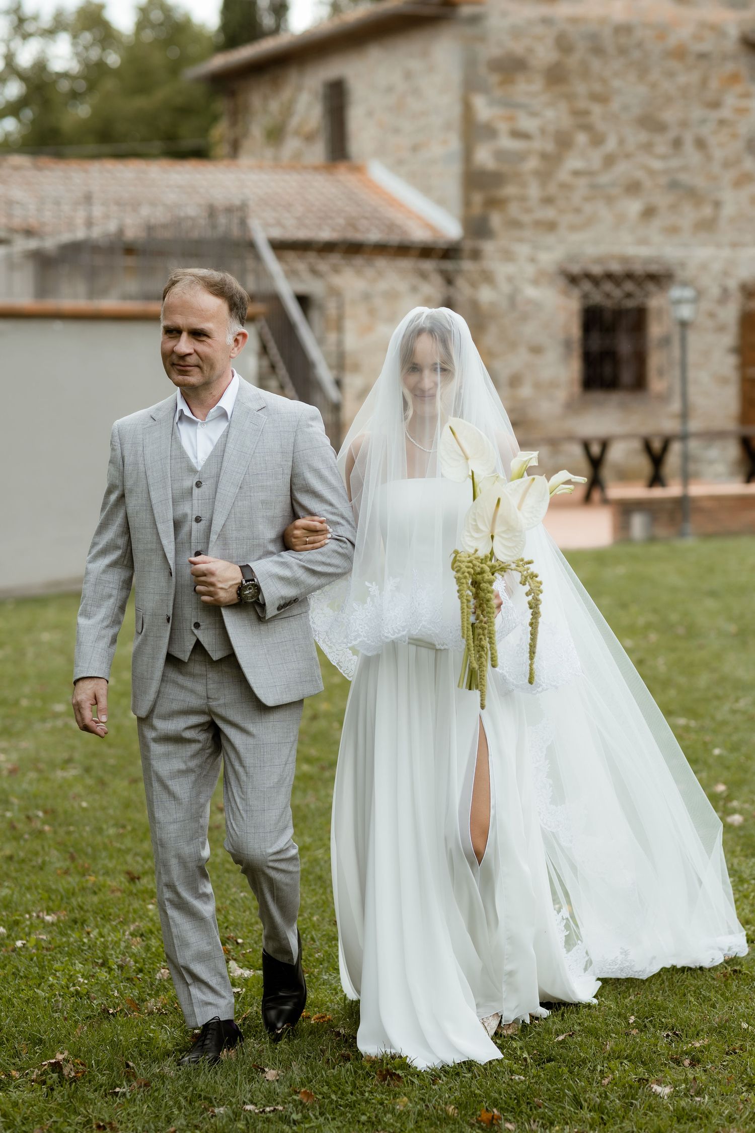 A couple in wedding attire walks together on grass with stone buildings in the background.