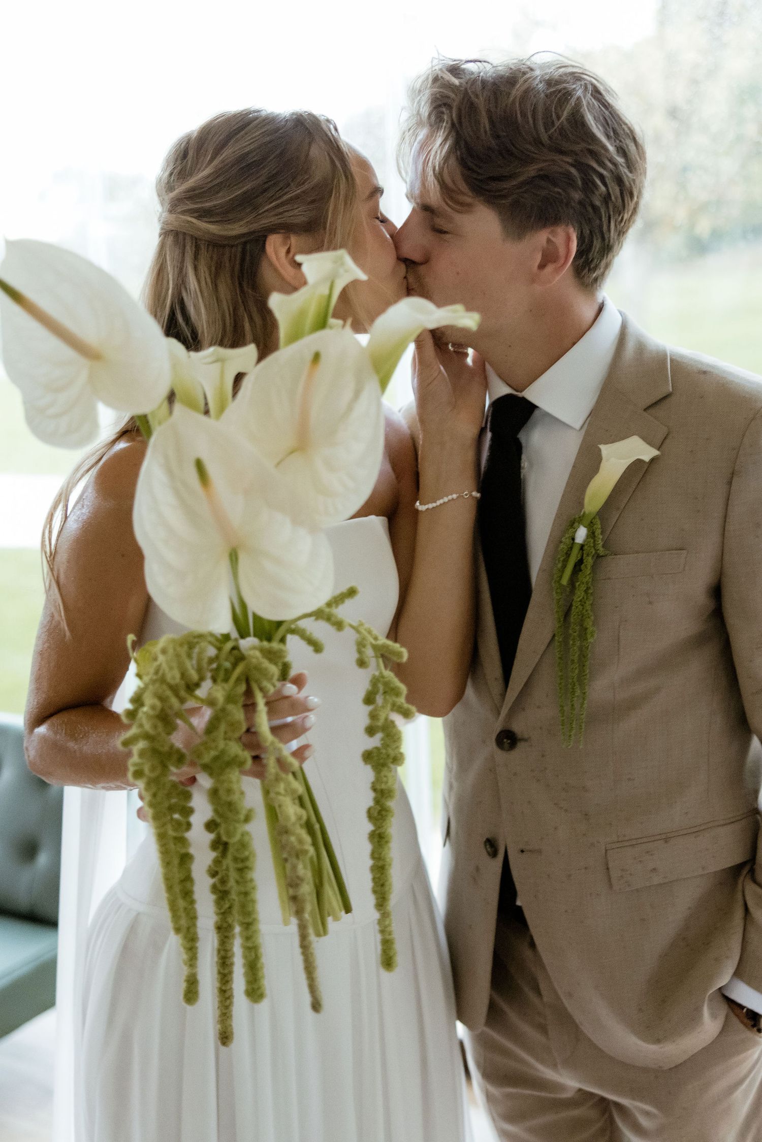 A romantic wedding couple shares intimate kisses while holding elegant white calla lily bouquets against a bright background.