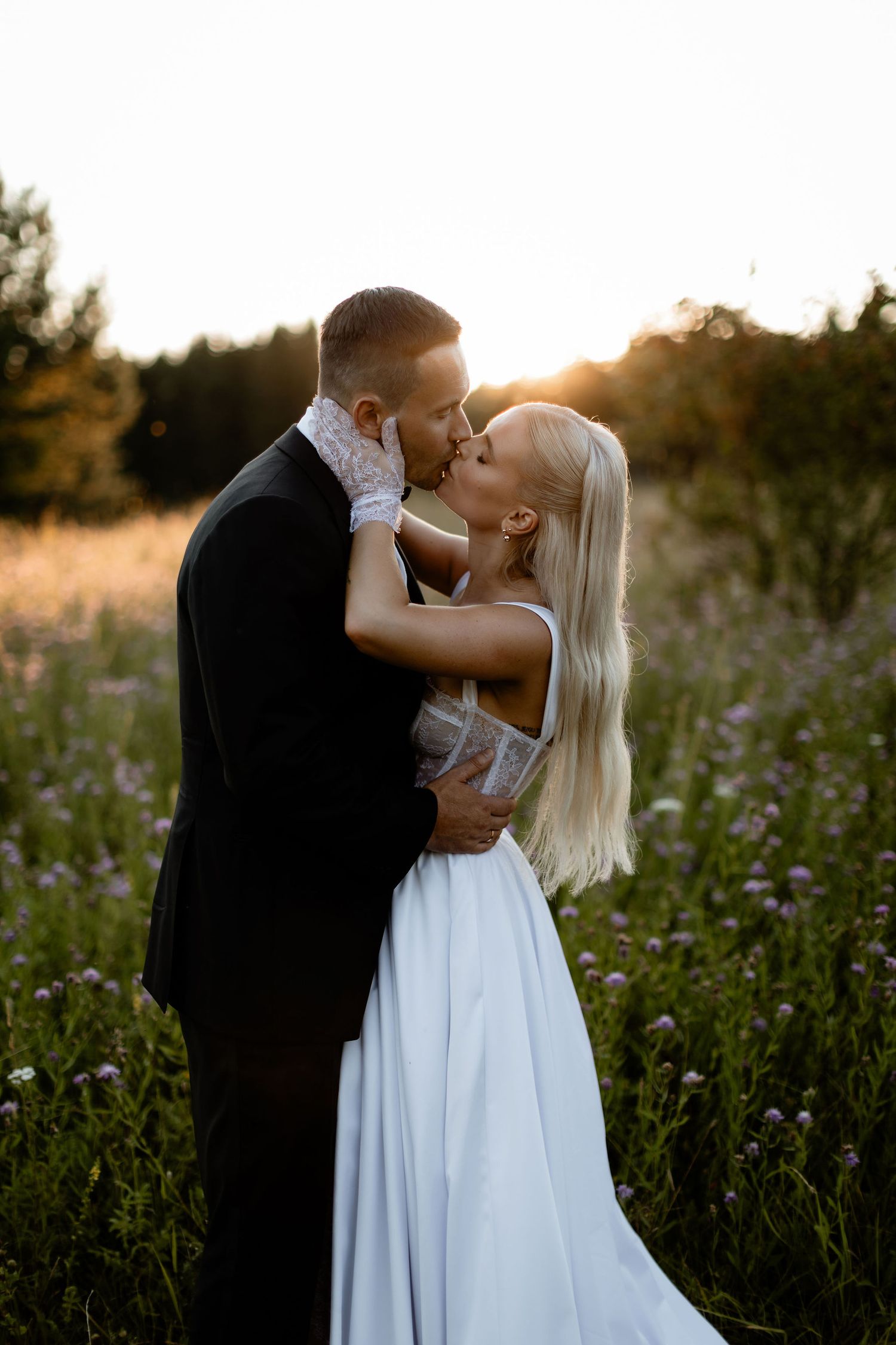 Silhouetted newlyweds share a kiss in a golden sunlit meadow during sunset.