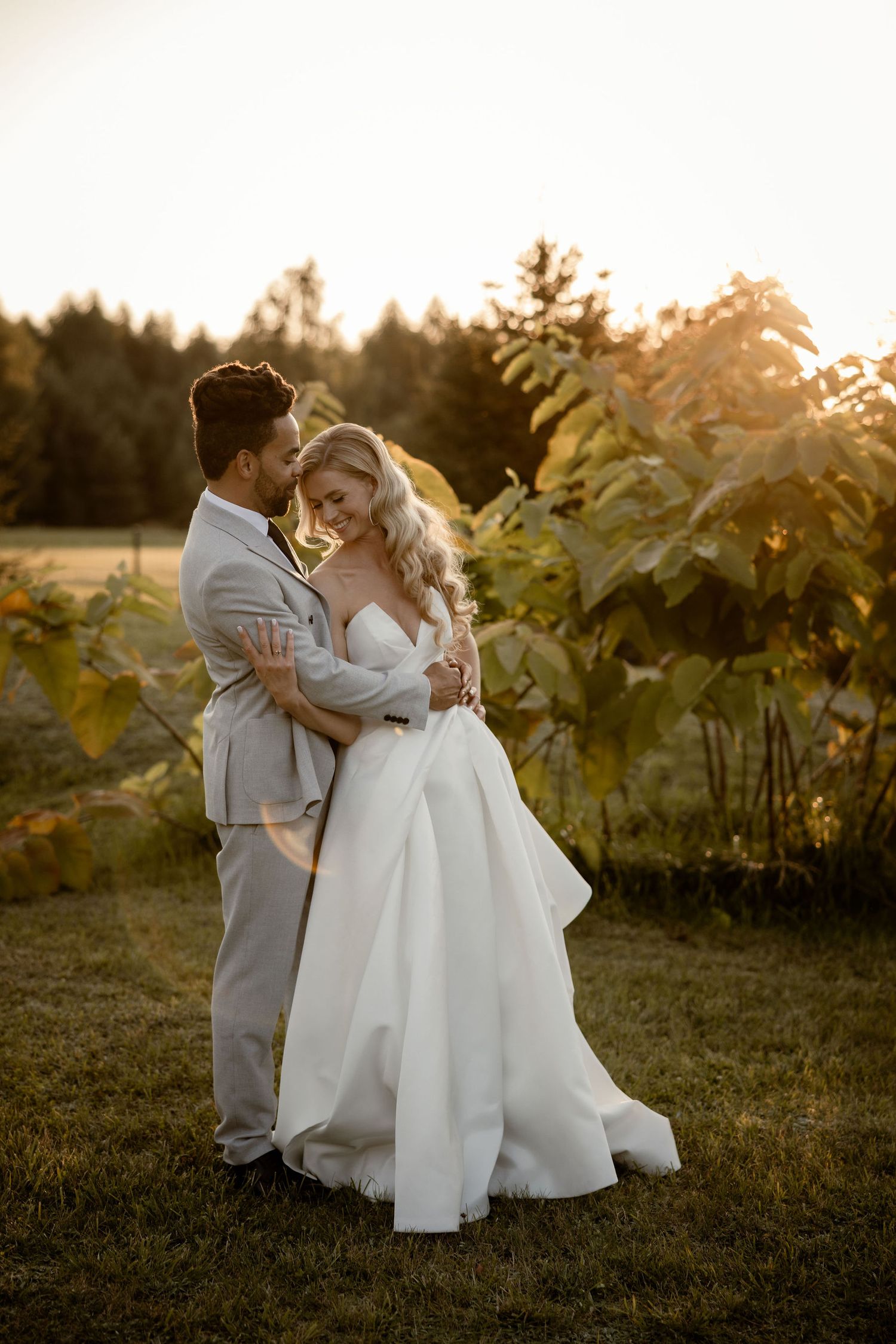 A couple shares a romantic embrace at sunset in a vineyard wearing wedding attire during golden hour.