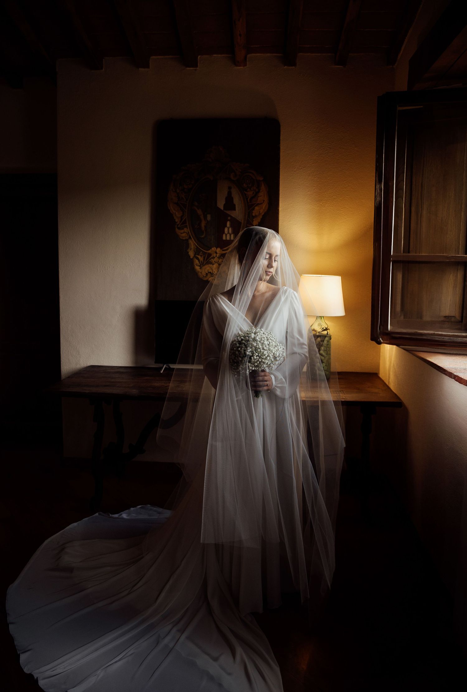 A bride in a flowing white wedding gown and long veil stands in a dimly lit room with warm lighting.