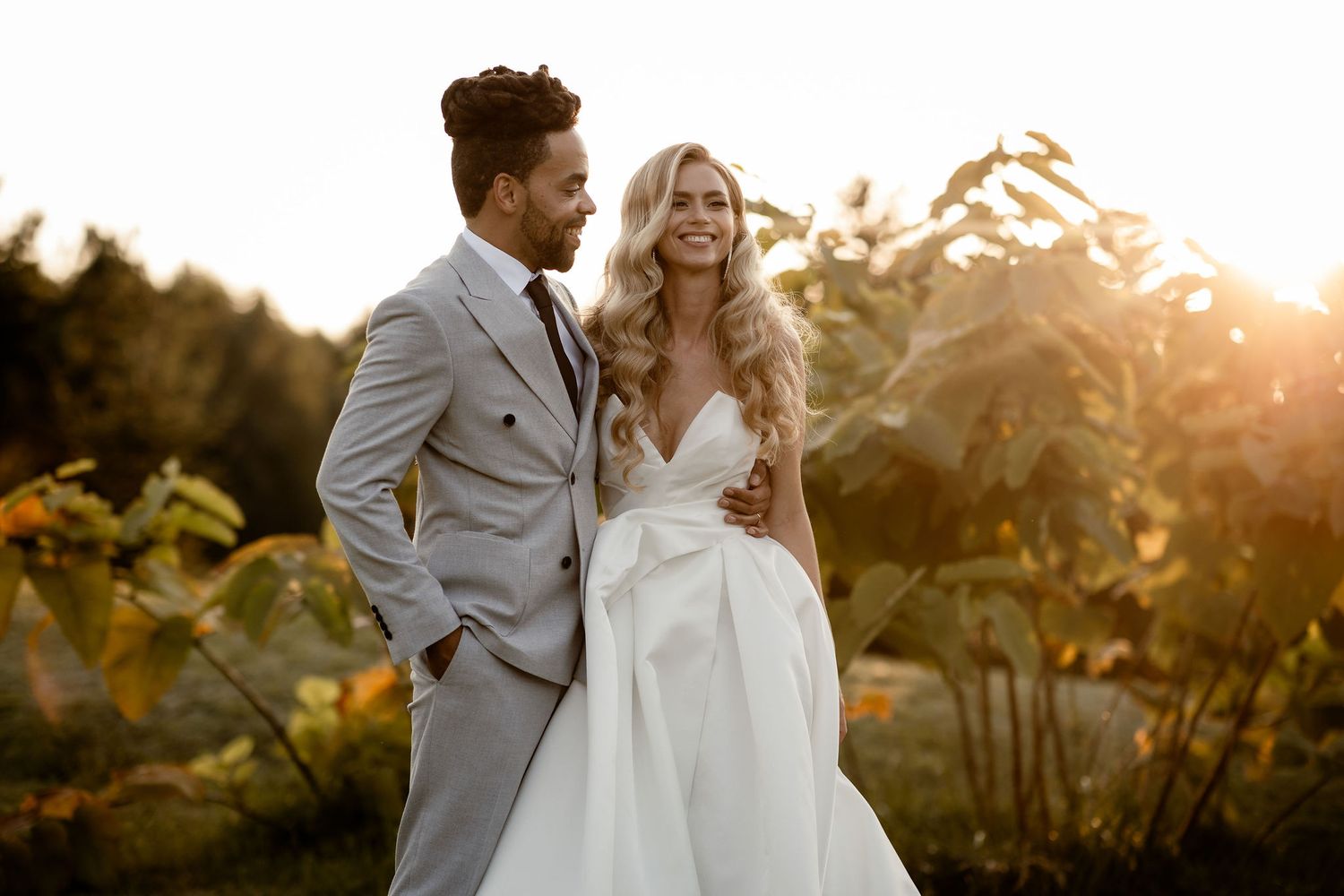 Couple in wedding attire pose in a sunlit garden during golden hour, with bride in white gown and groom in gray suit.