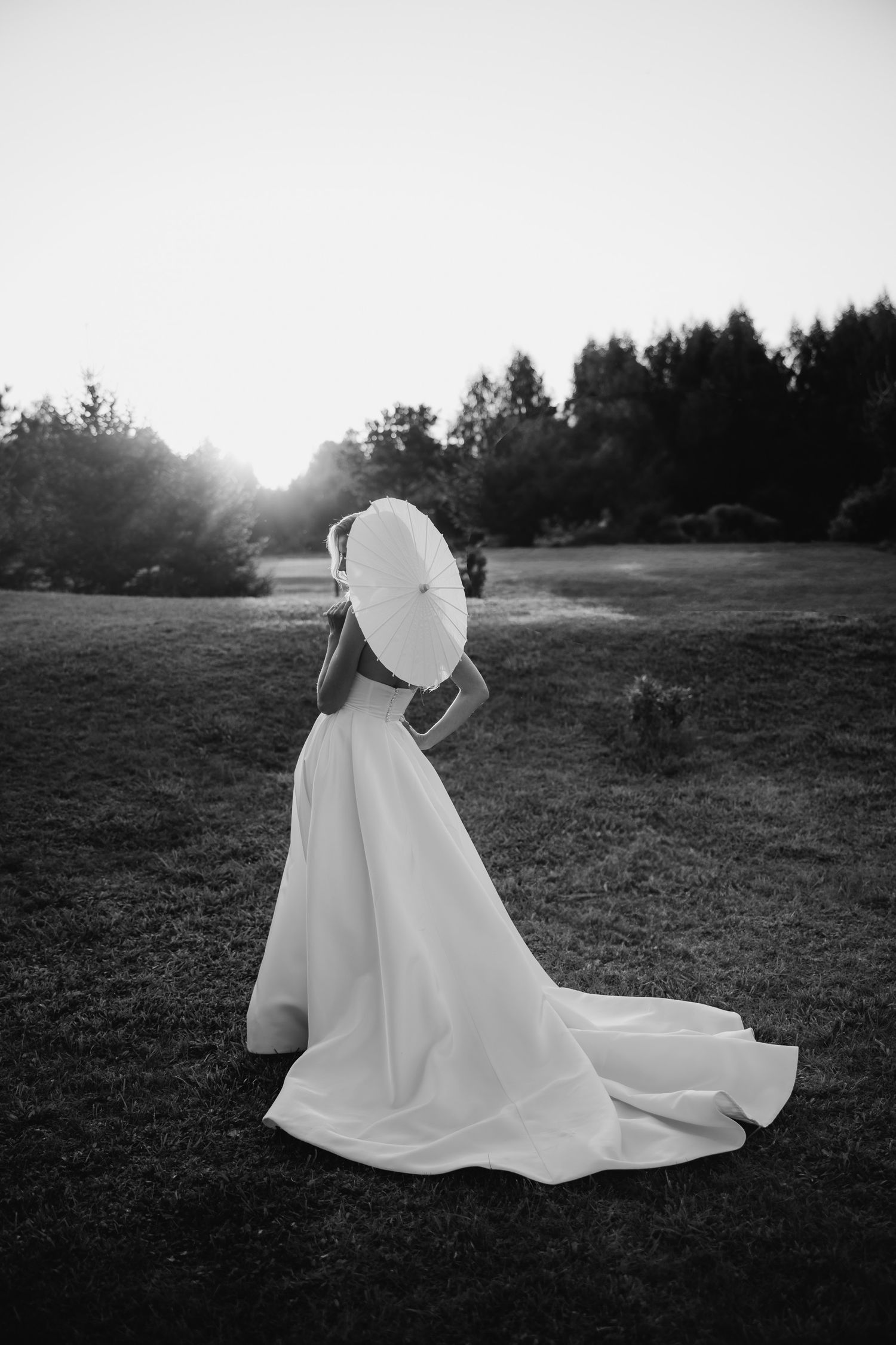 Black and white photo of person in elegant white wedding gown holding parasol outdoors at sunset in field.