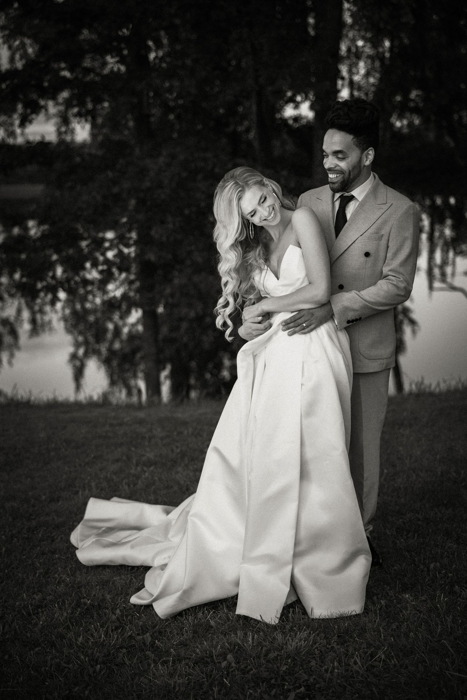 Black and white wedding photo of a couple embracing outdoors by a lake with trees in the background.
