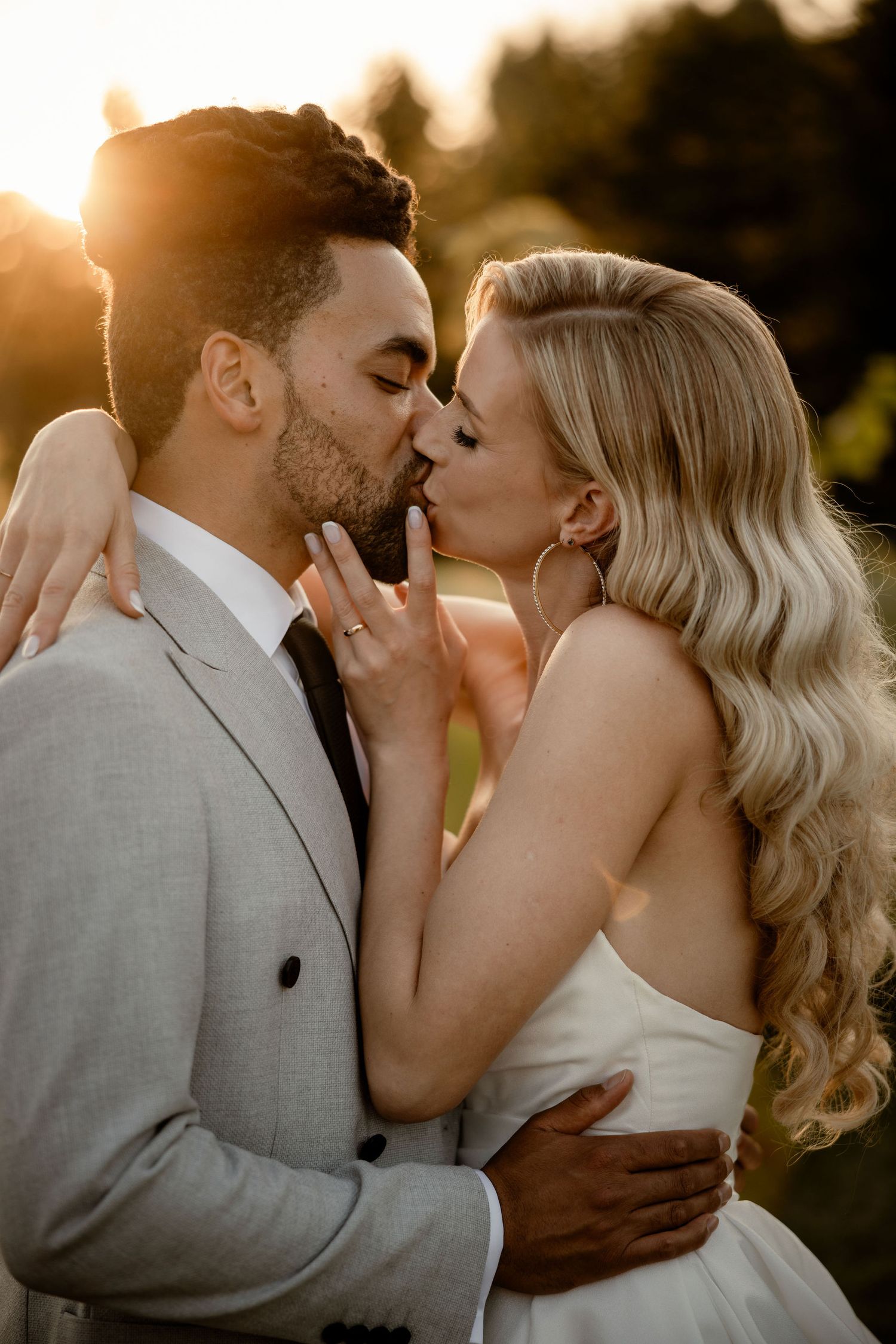 Romantic sunset wedding kiss between a couple in grey suit and white dress with golden backlighting through trees.