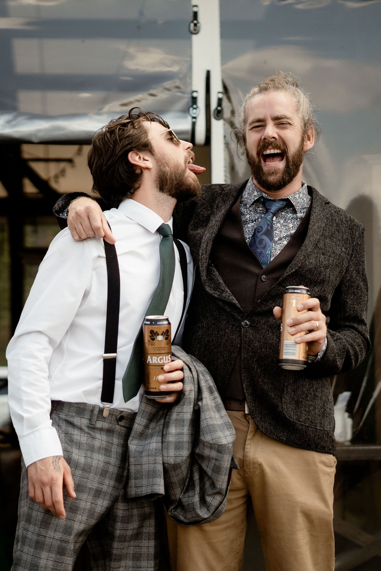 Two bearded friends in vintage attire sharing laughs while holding coffee cups outdoors.