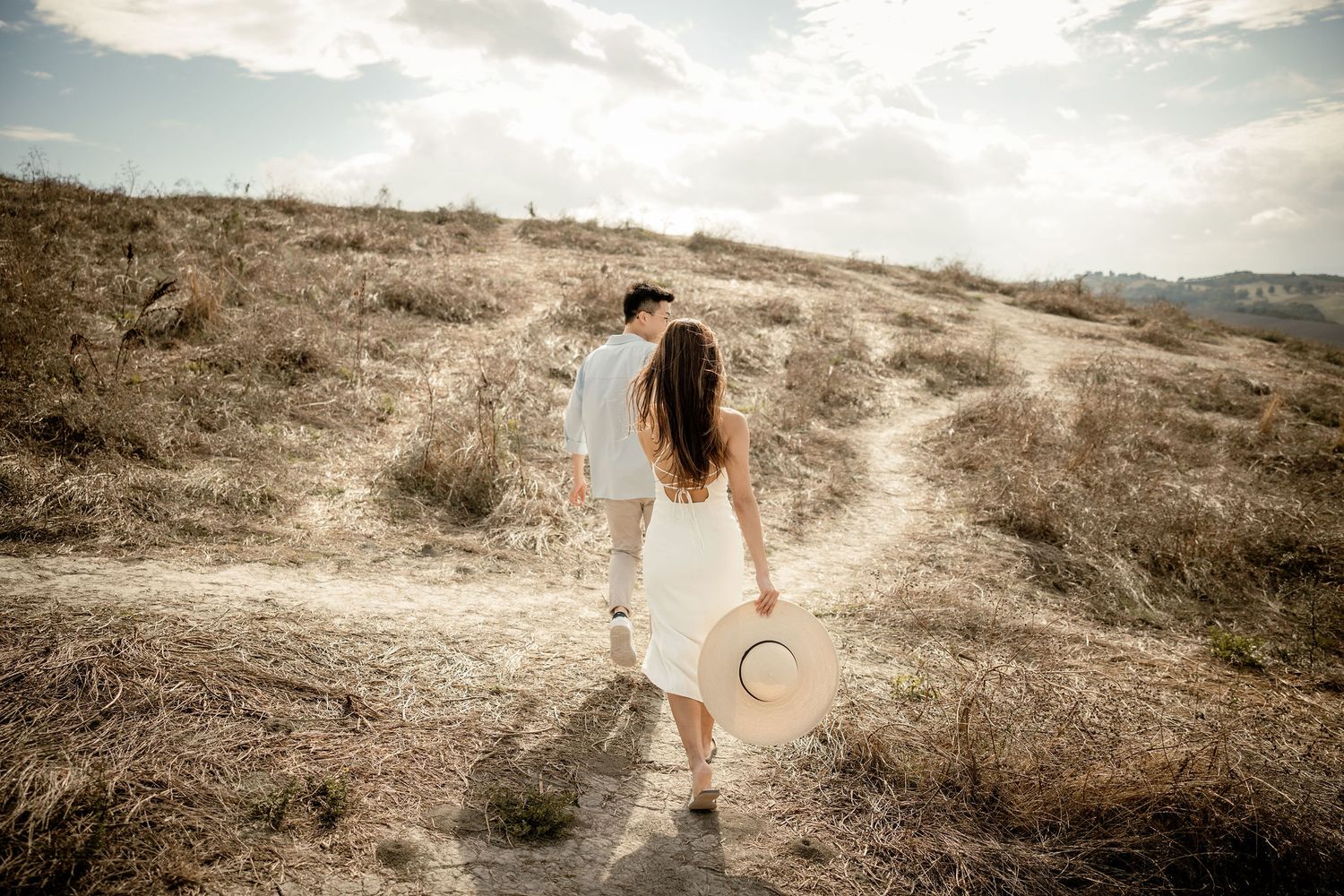 Woman in white dress walking on trail holding straw hat during sunset.