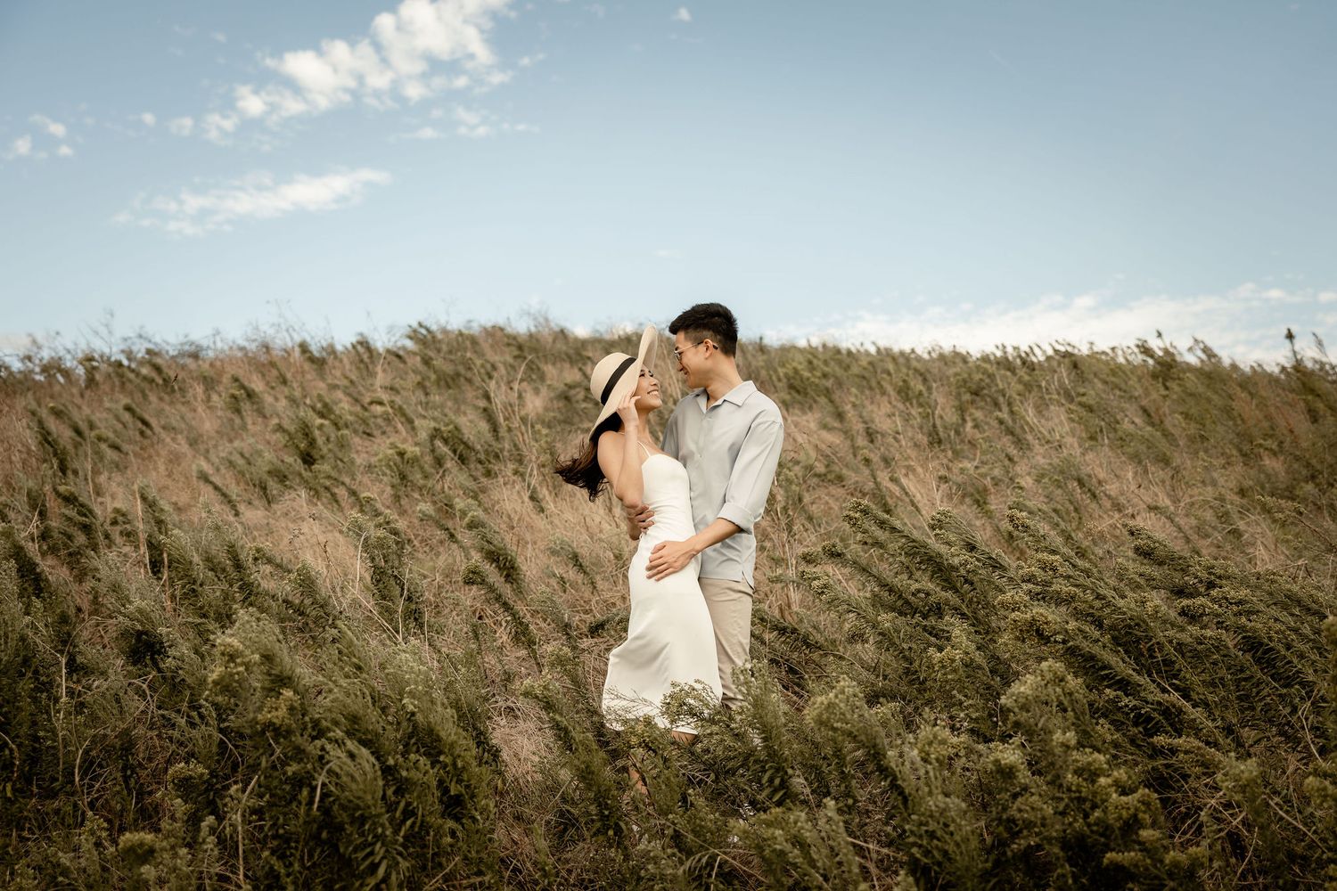 A couple embraces in a windswept field of grass under blue skies.