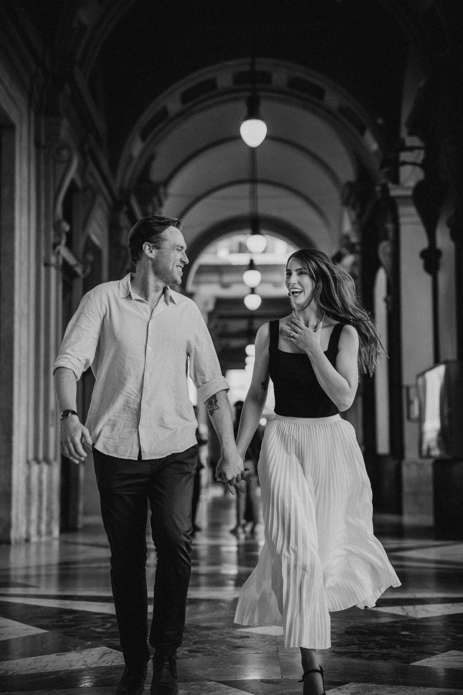 A couple in elegant attire running and laughing together through an ornate arched hallway lit by pendant lights.