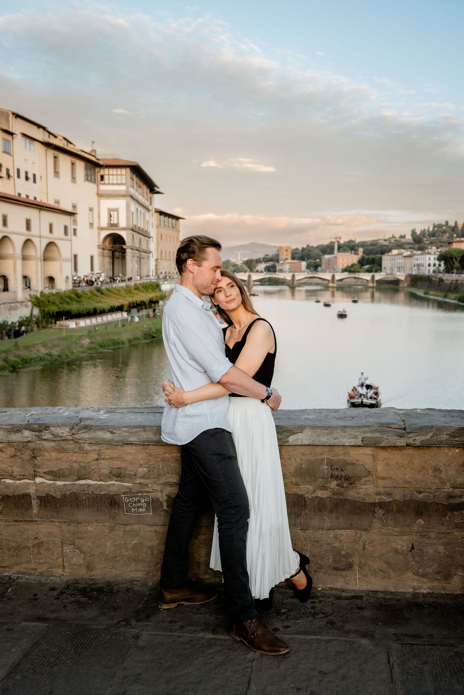 A couple embraces on a stone wall overlooking the Arno River with historic Florentine architecture in the background.