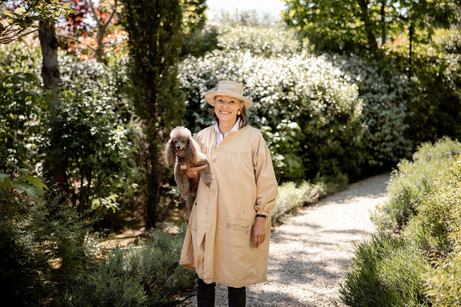 Someone wearing a beige coat and sun hat stands on a garden path with blooming white flowers in the background.