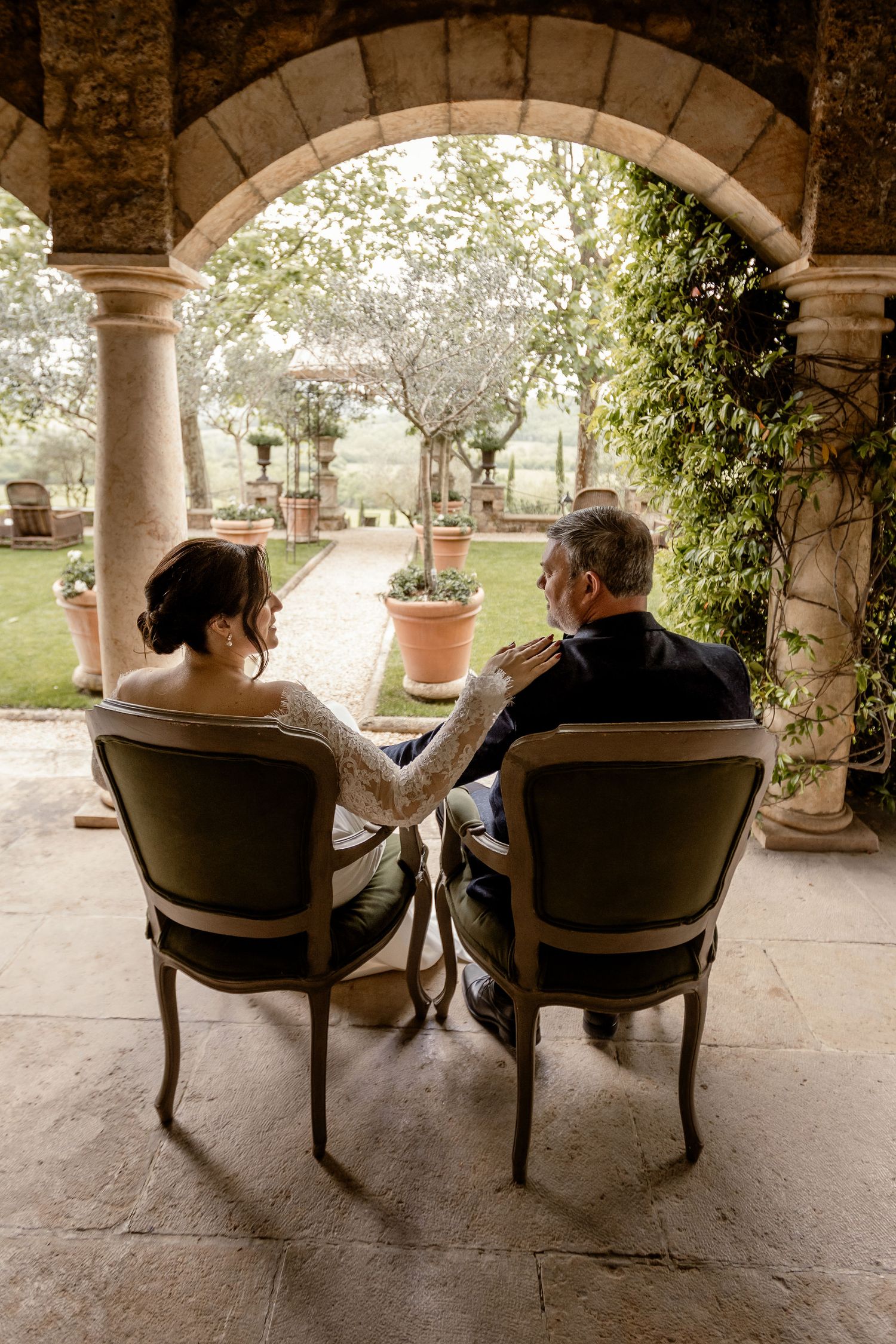 Couple sits together under stone archway overlooking garden patio with potted plants and trees.
