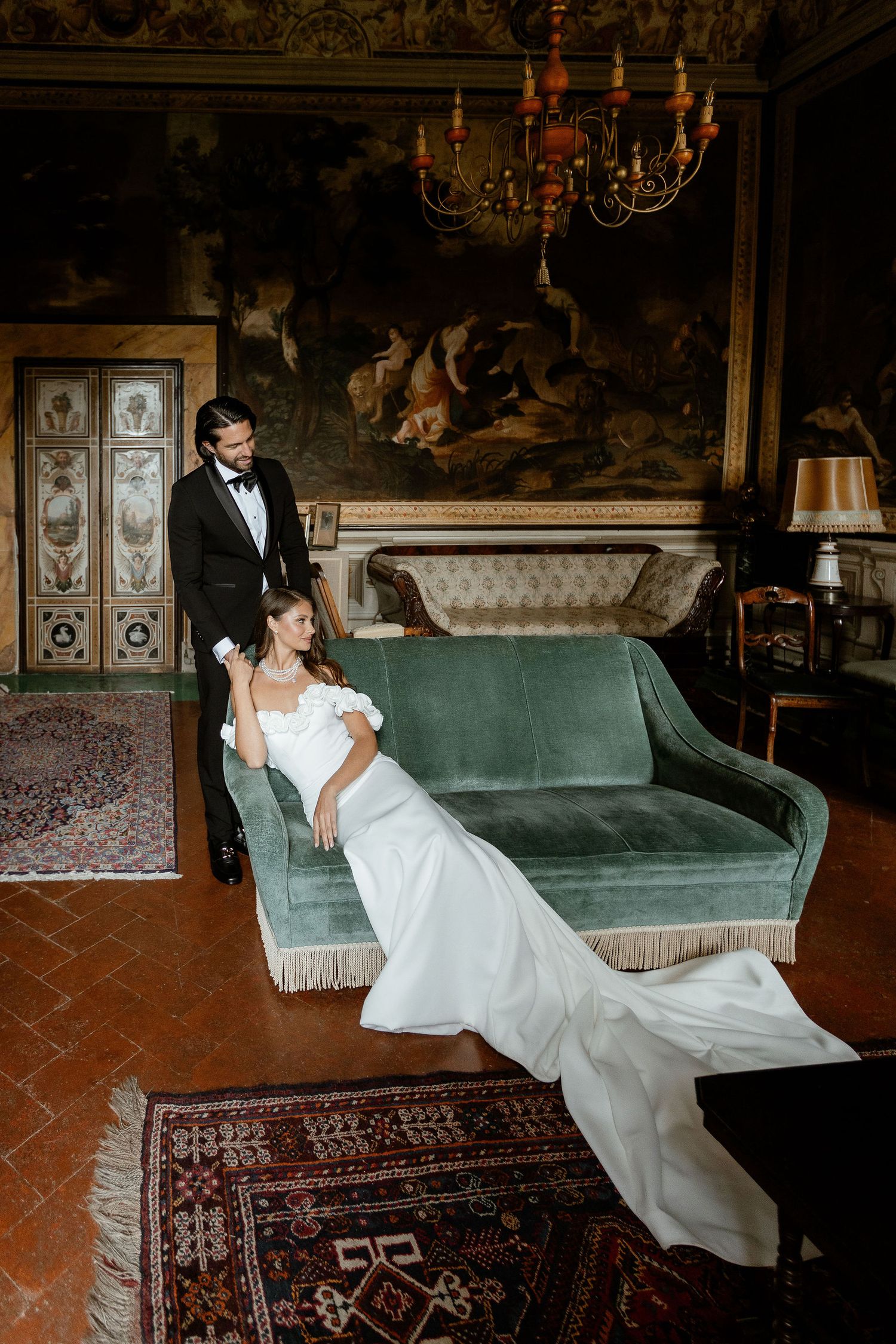 A grand vintage interior with an emerald green velvet couch, ornate chandelier and Persian rug in an elegant wedding setting.