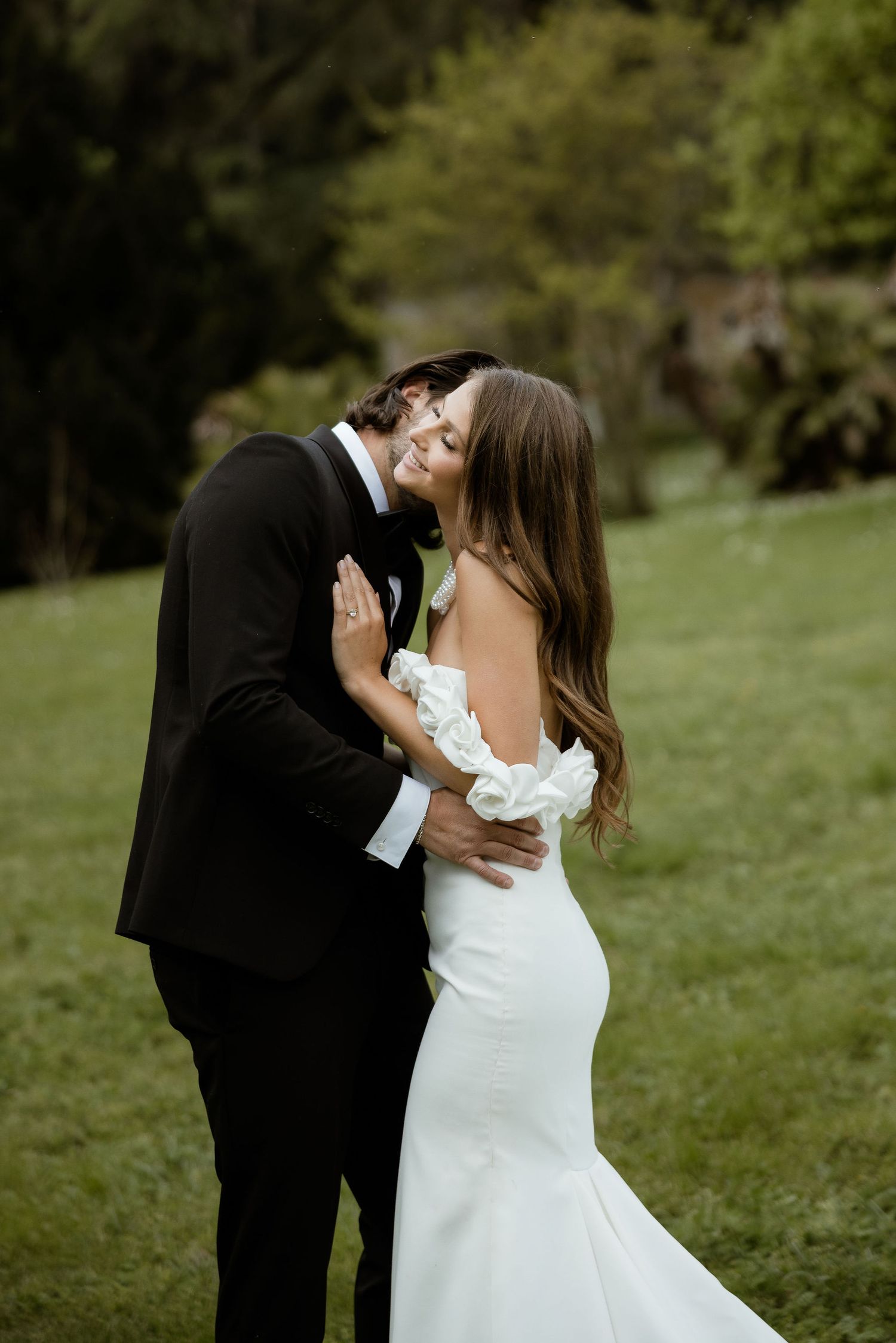 A romantic outdoor wedding photo shows a couple in formal attire embracing on a lush green lawn.