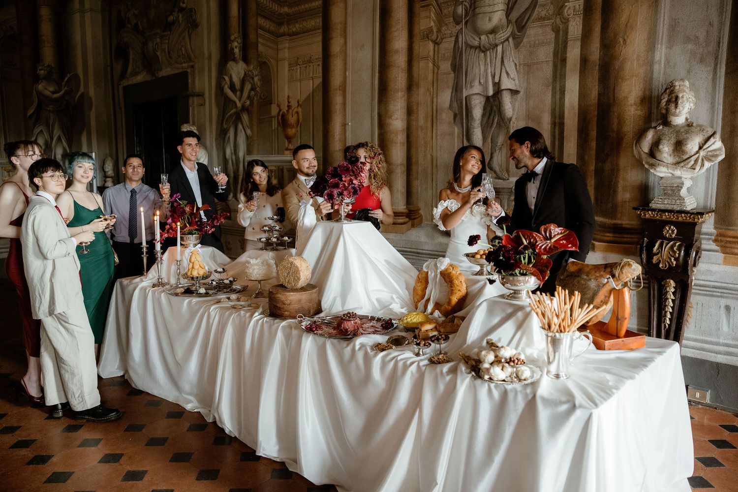 Elegant reception buffet table with white linens and candles set in an ornate historic ballroom with guests gathered around.