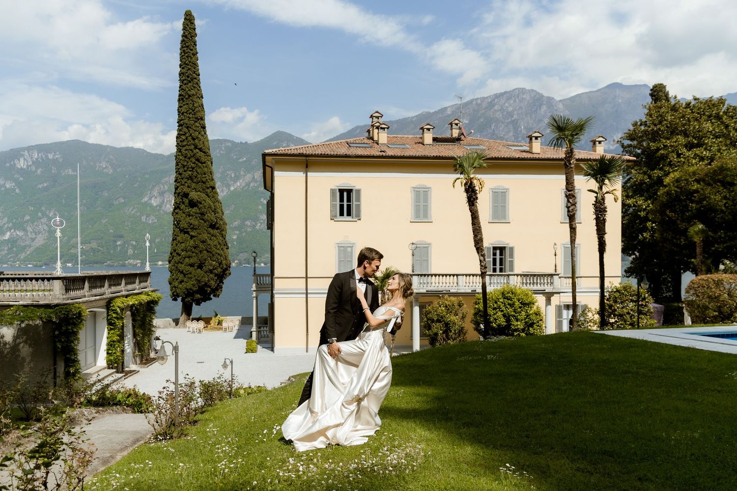 Bride and groom share romantic dip kiss on lush lawn of elegant Italian villa with lake and mountains backdrop.