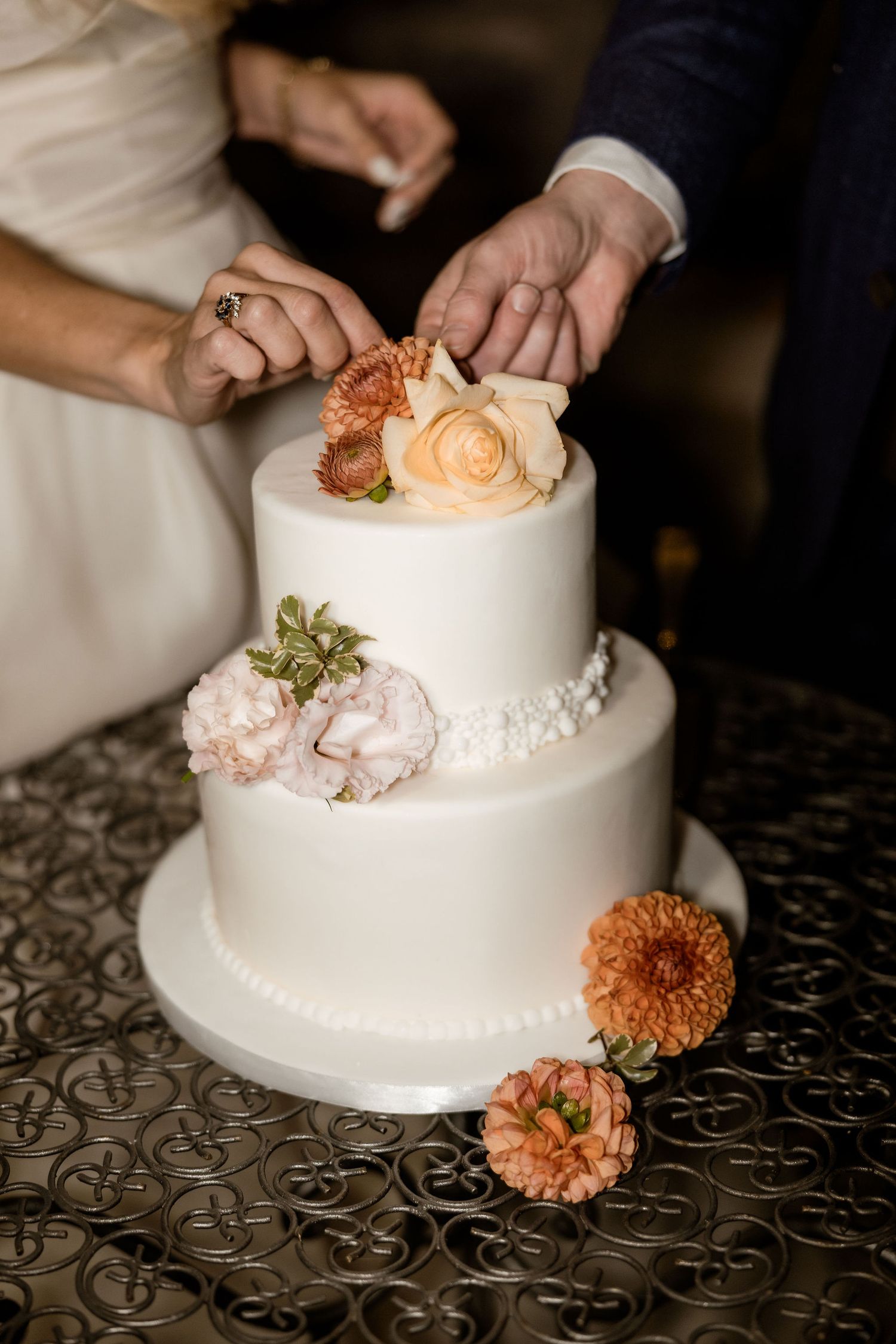 Wedding cake decorator carefully arranges fresh peach and coral flowers on a classic white tiered cake.