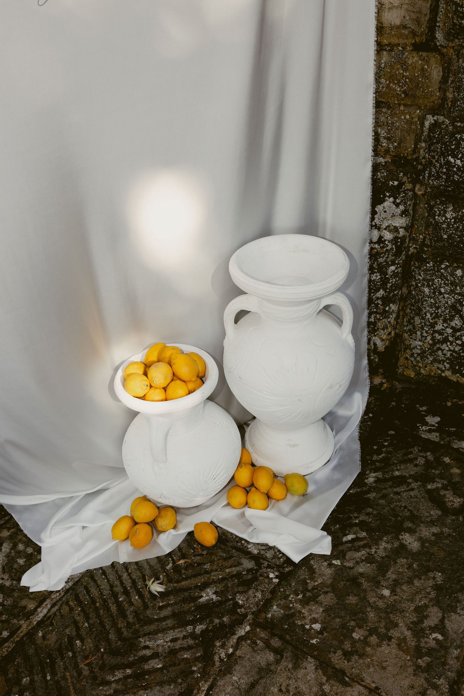 White ceramic vases and bowls with fresh yellow kumquats arranged on white fabric against a textured dark wall.