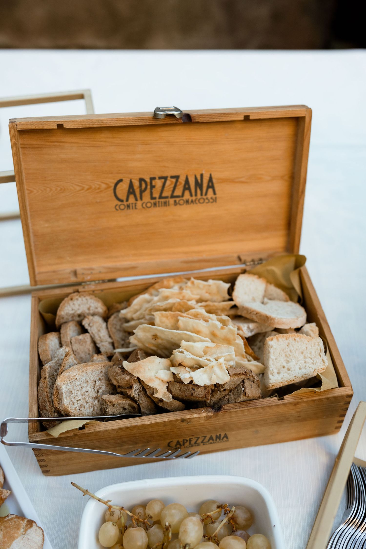 Rustic wooden bread box labeled Capezzana displaying sliced artisanal bread and serving utensils.