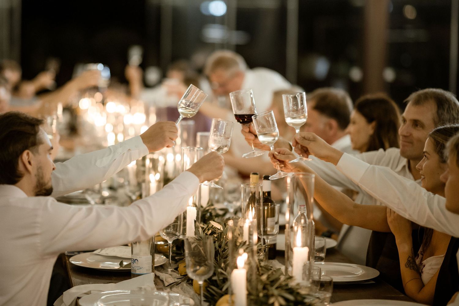 Guests raise wine glasses in a toast during an elegant candlelit dinner party at a long table.