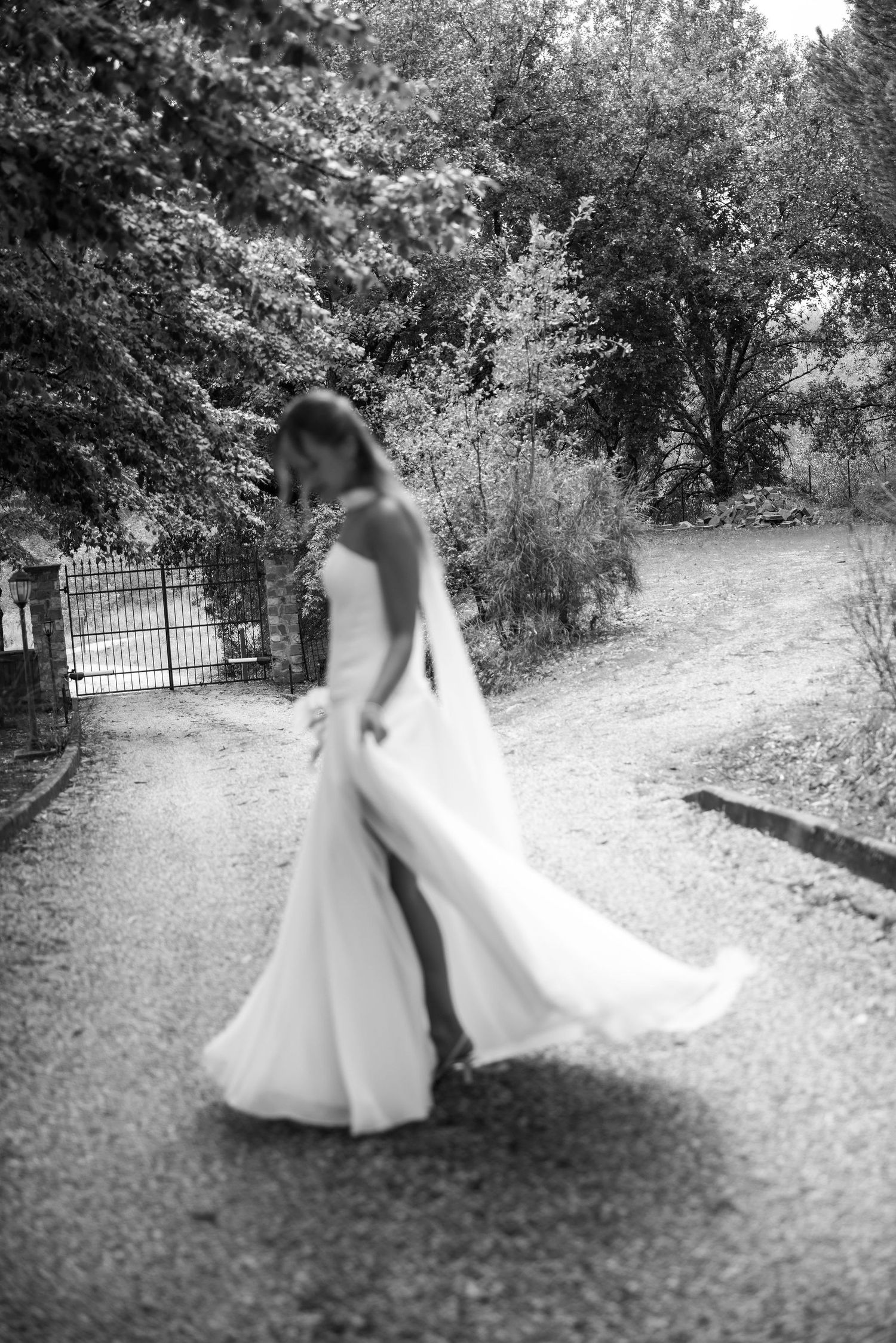 A bride in a flowing white wedding dress twirls gracefully along a tree-lined garden path in black and white photography.