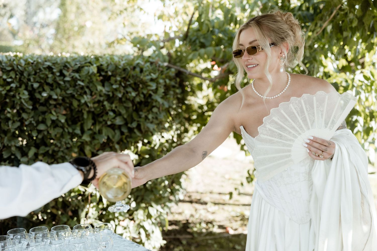 A bride in white dress holding a decorative fan during an outdoor garden celebration.