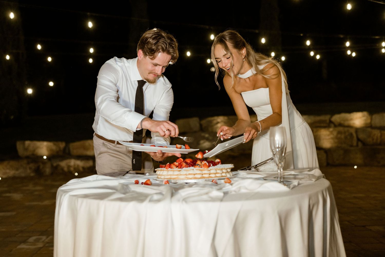 Couple sits at a round table with string lights overhead as they share a slice of red velvet cake together.