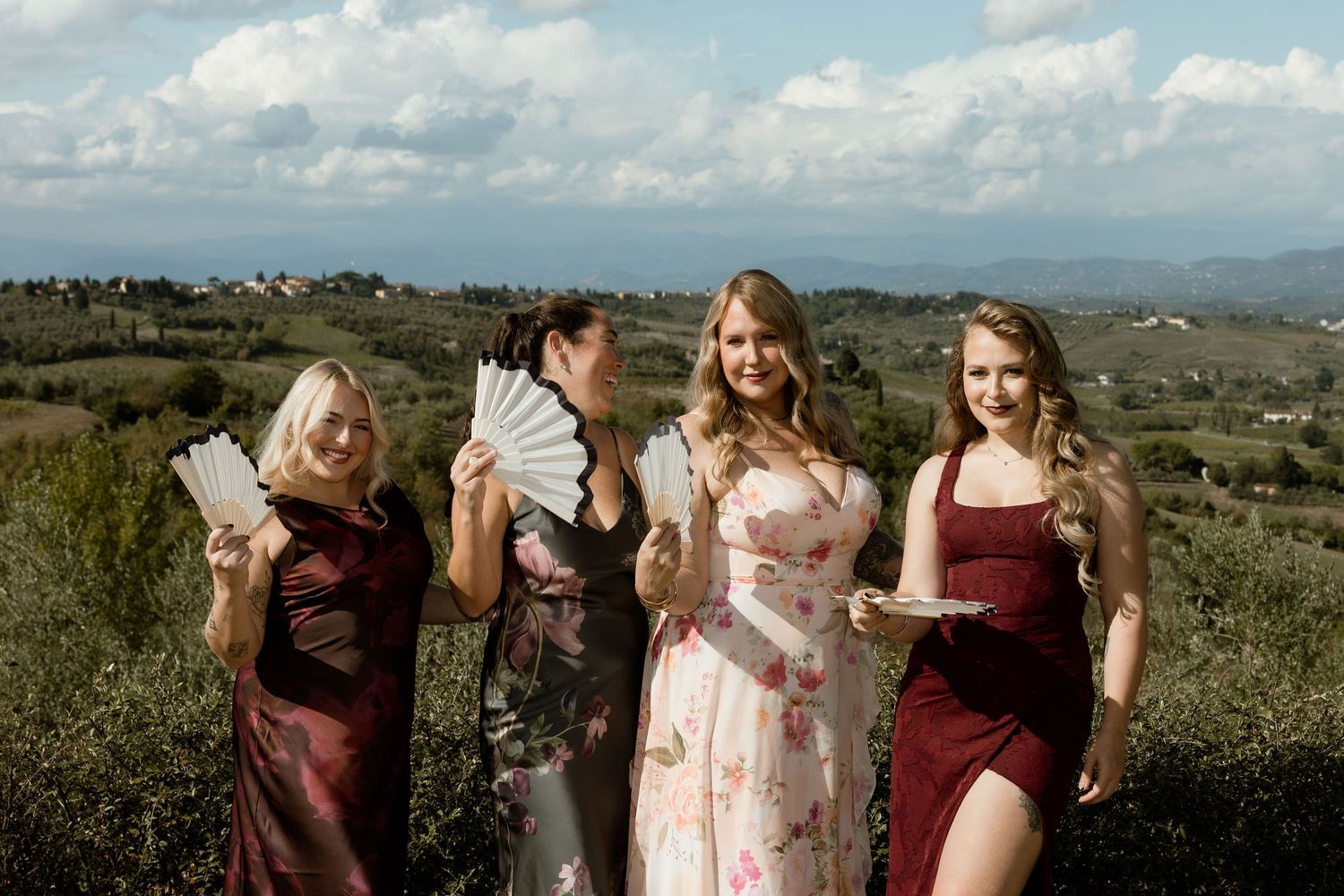 Bridesmaids in burgundy and pink dresses gather in a scenic field under cloudy skies during a wedding photoshoot.