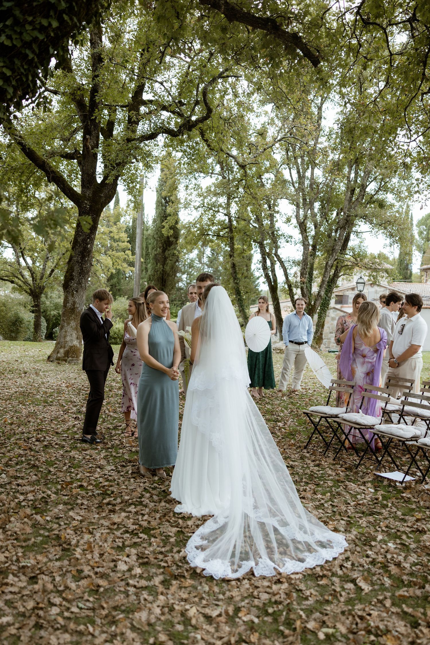 An outdoor wedding ceremony in a wooded area with guests gathered around folding chairs and tall trees overhead.