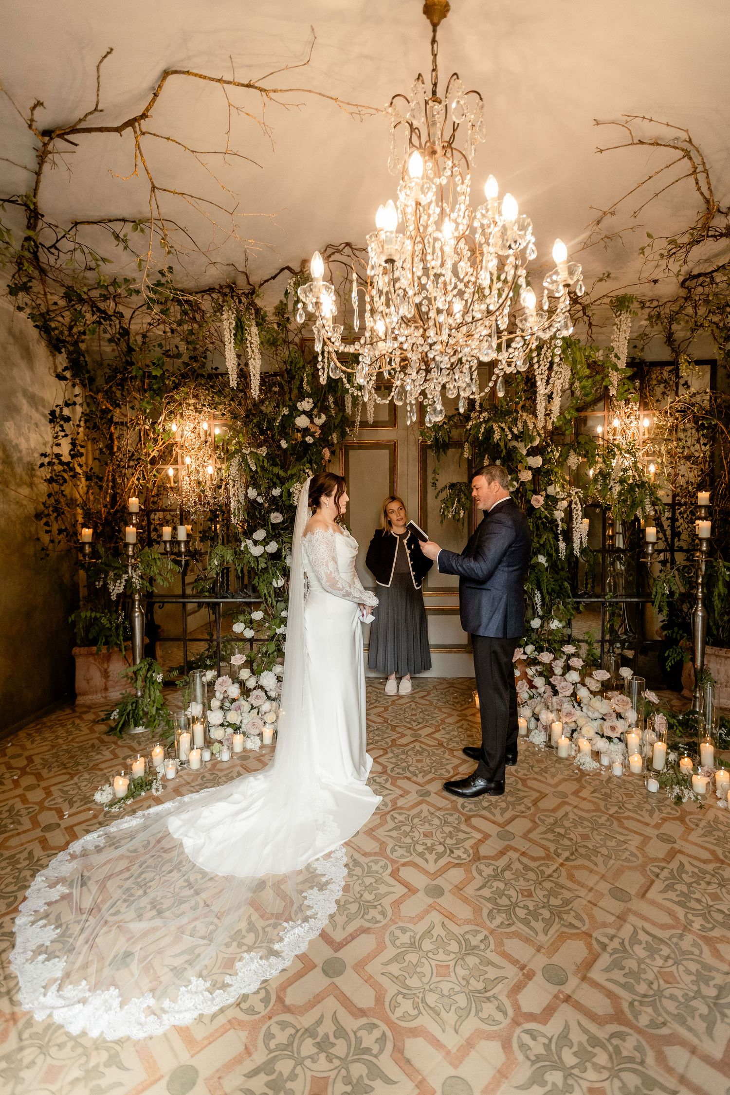 Elegantly decorated indoor wedding ceremony with crystal chandelier, floral arrangements and candles on ornate tiled floor.
