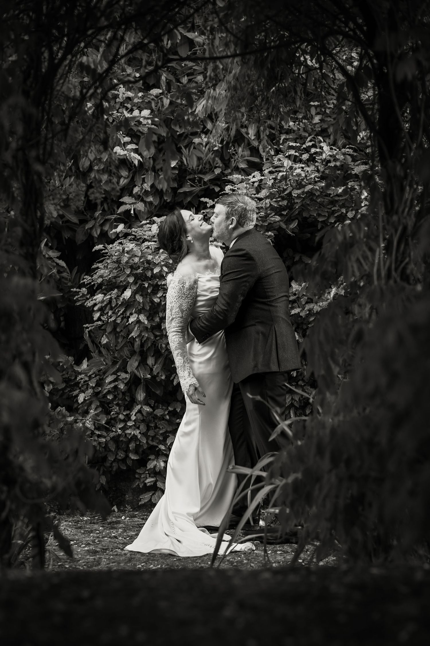 Black and white artistic wedding photograph of a couple embracing on a garden path surrounded by lush foliage.