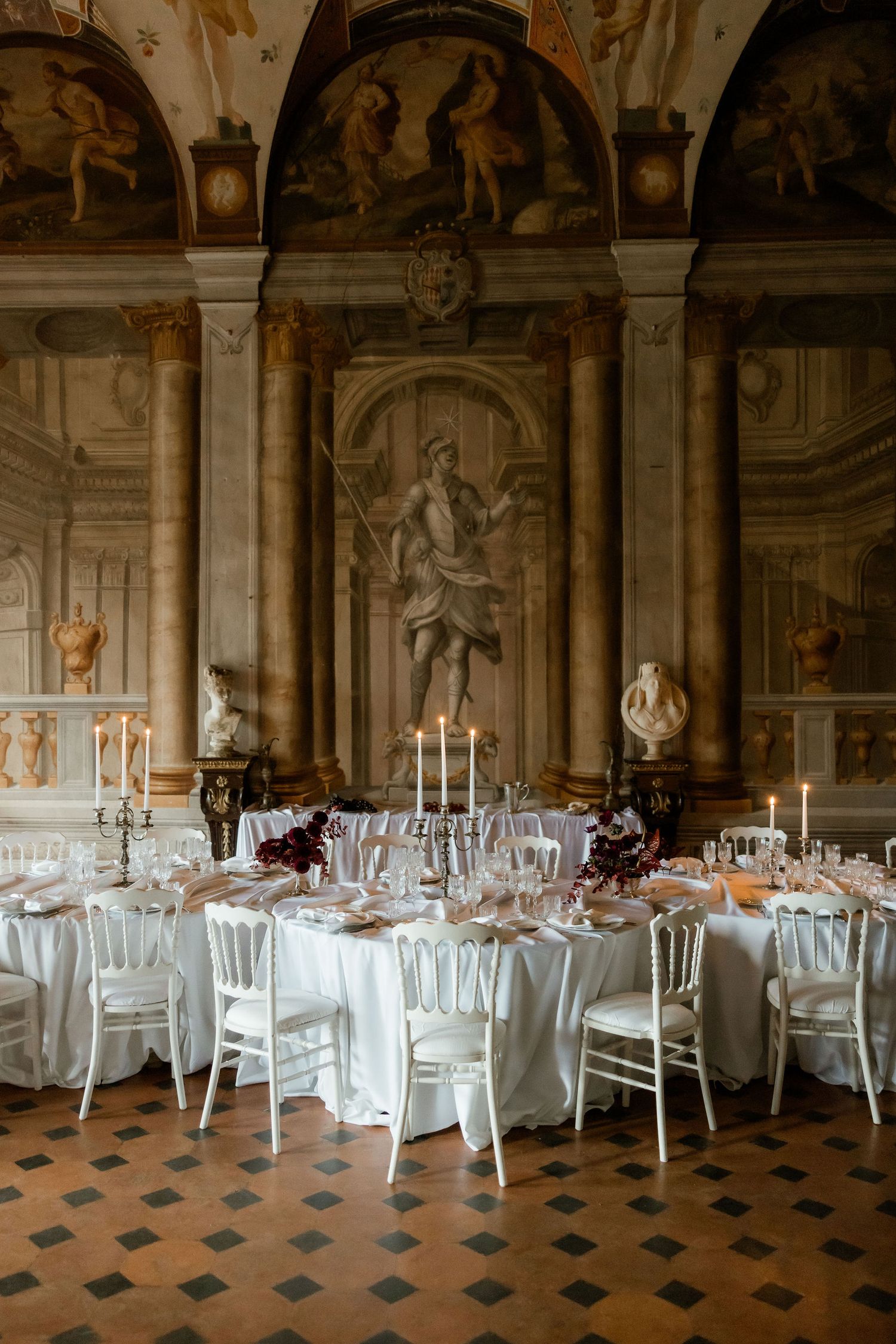 Elegant wedding reception tables set with white linens and chairs in an ornate baroque hall with classical frescoes and statues.